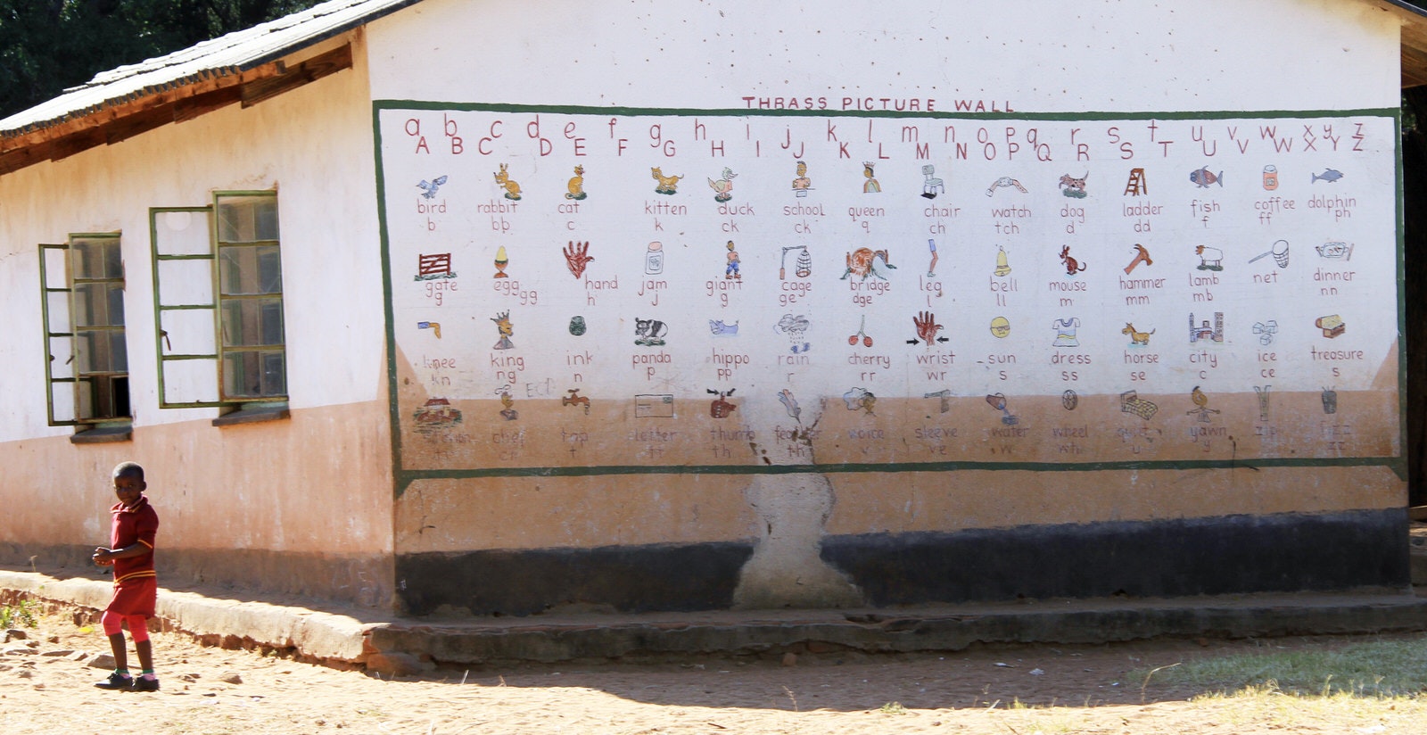 A young child stands next to a single-room school with two windows; painted on the end exterior wall is the alphabet and images for each letter