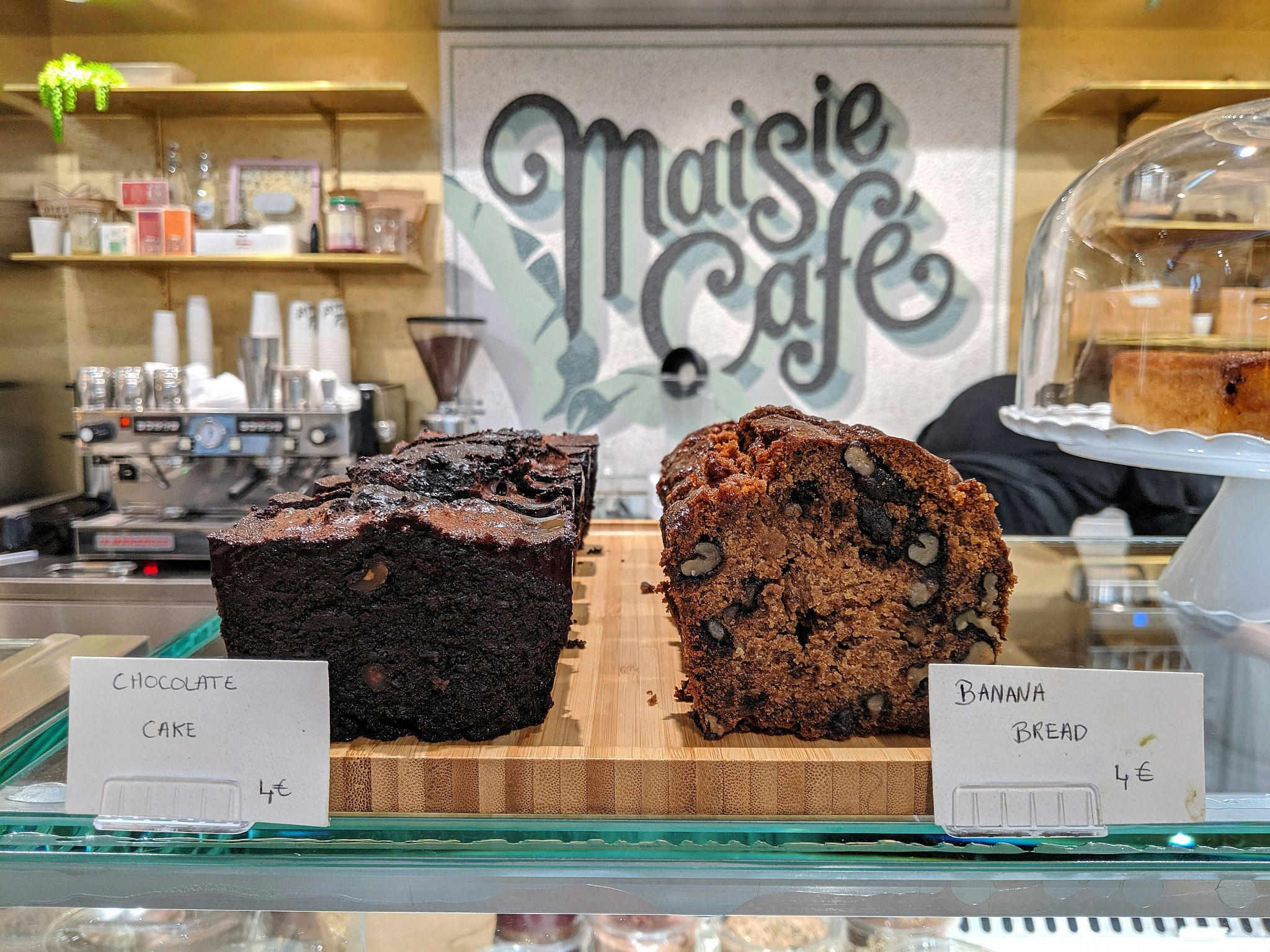 A chocolate cake and a banana cake on a wooden chopping board on a glass shop counter.