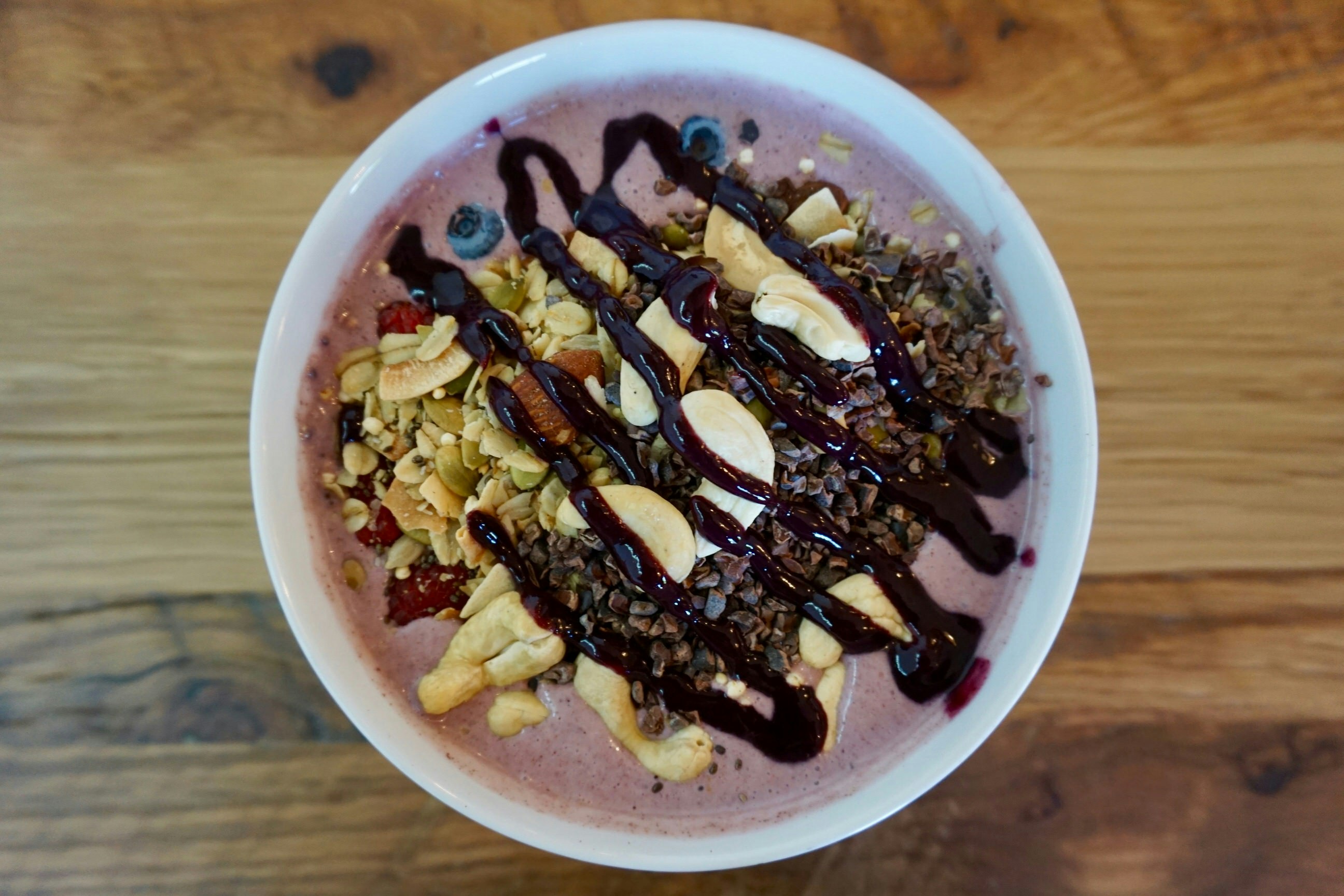 A white bowl filled with pink yogurt, nuts, granola and fresh fruits such as blueberries and raspberries, all topped with a generous serving of deep red syrup.