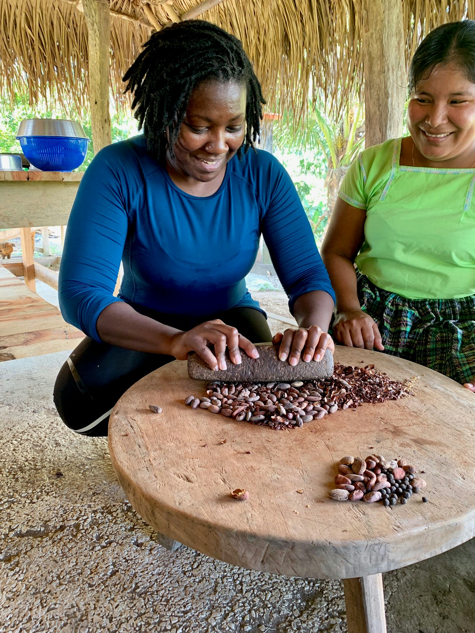 A Mayan woman shows another woman how to crush cacao beans with a volcanic rock pestle