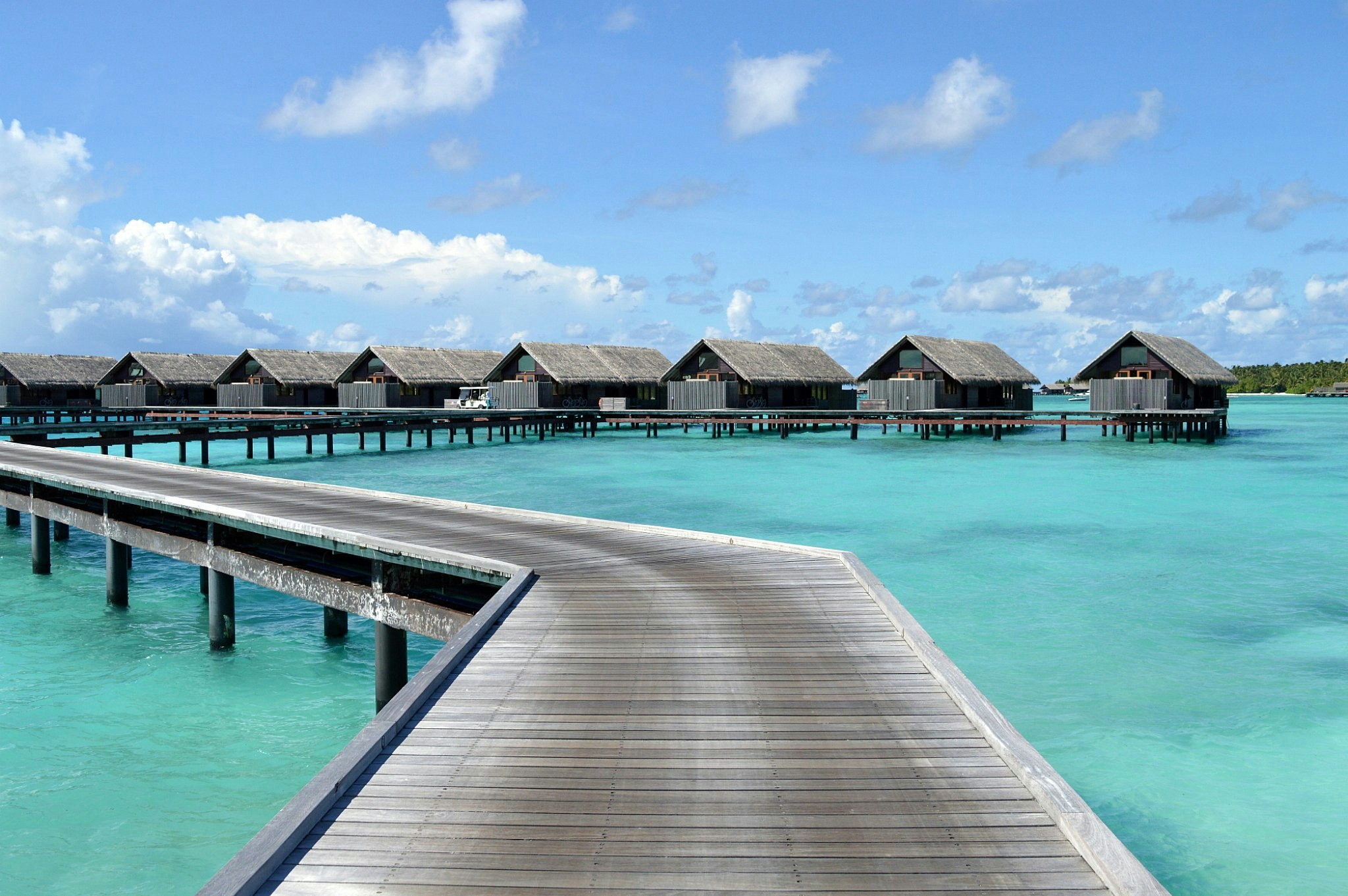 A wooden boardwalk stretches out towards a row of wooden overwater villas above clear blue water.