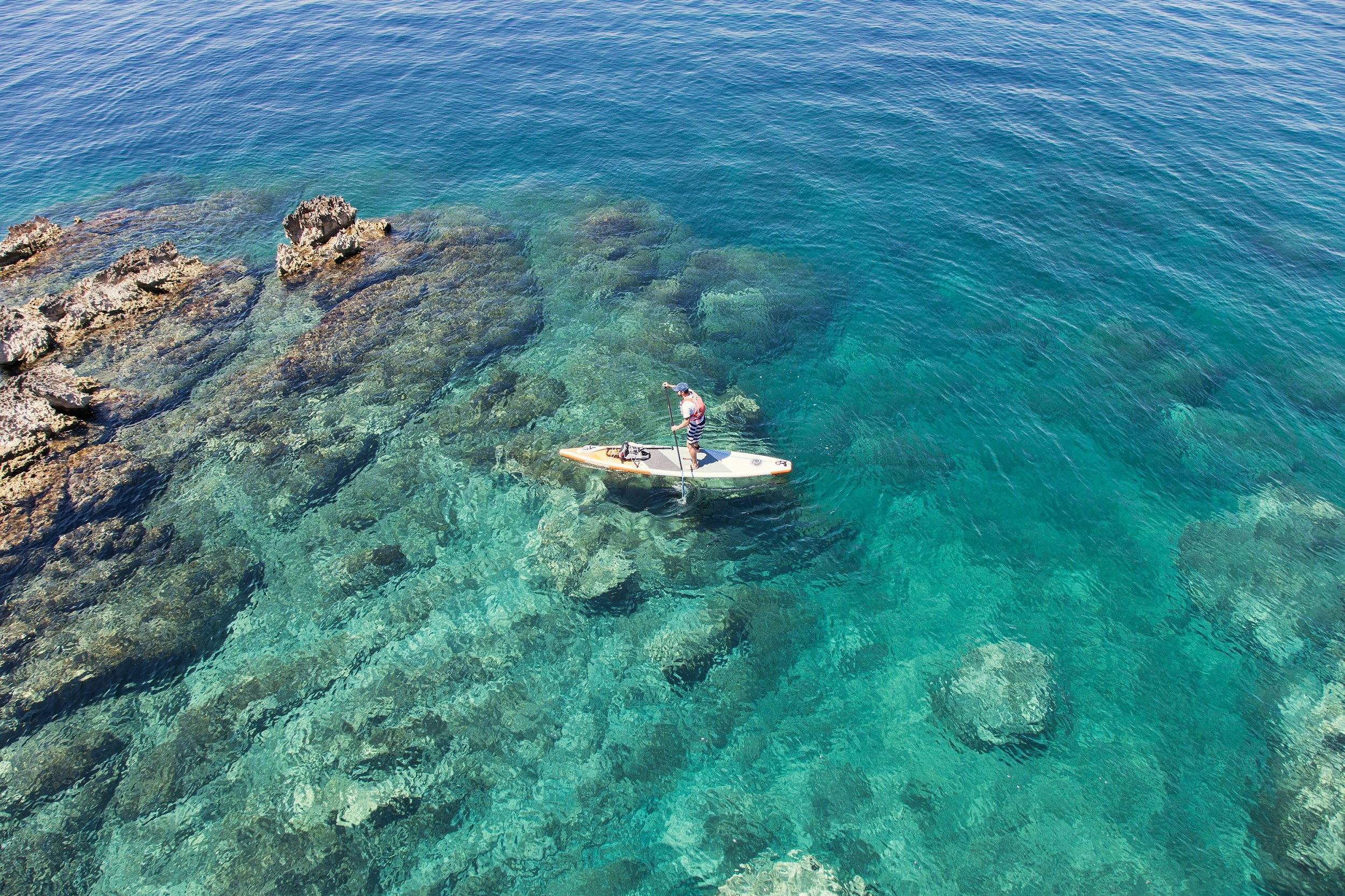 Shot from above, a man paddles an SUP along a rocky shore atop turquoise waters.