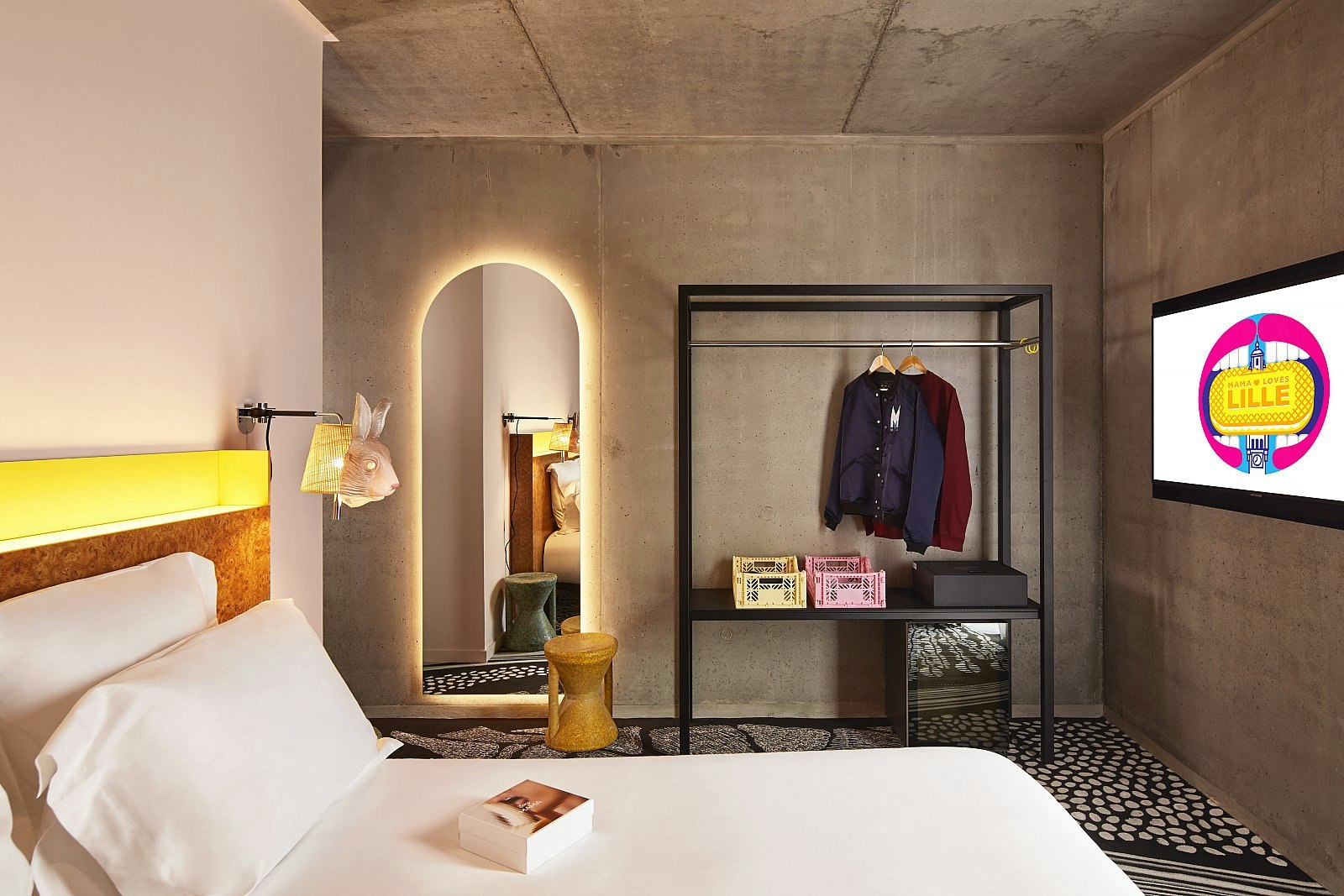 A bedroom at the Mama Shelter Lille hotel: walls have an exposed concrete finish; the bed has white linen and a wooden headboard; there is a plastic rabbit mask over the bedside light.
