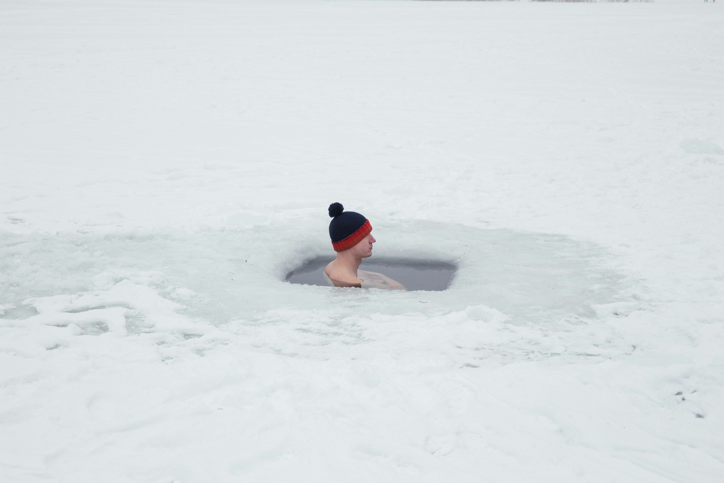 A man wearing a black and red bobble hat sits in an ice hole.