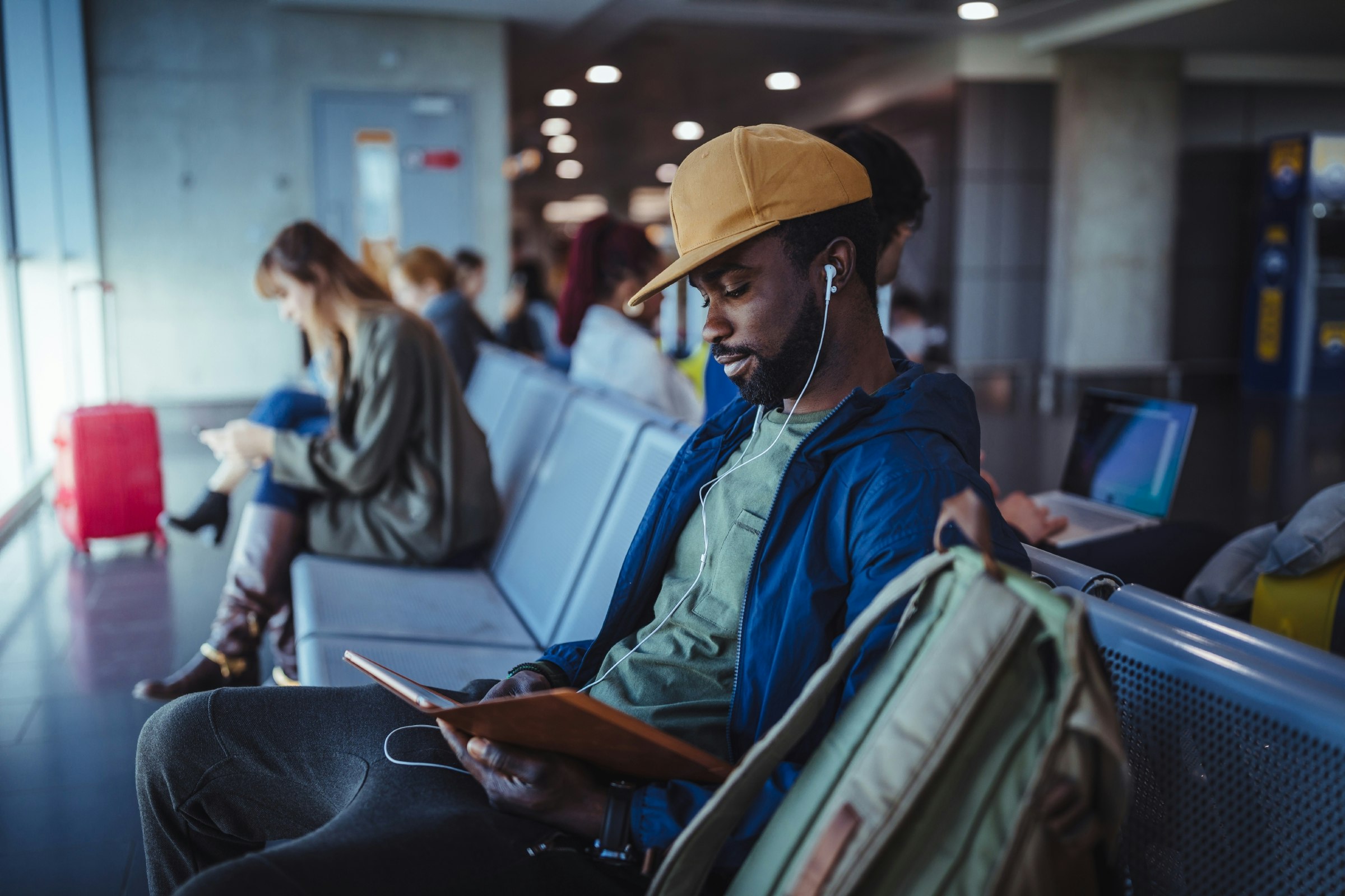 A man listens to music while reading an ebook in an airport departure hall. His bag is next to him and fellow passengers are sitting around him.