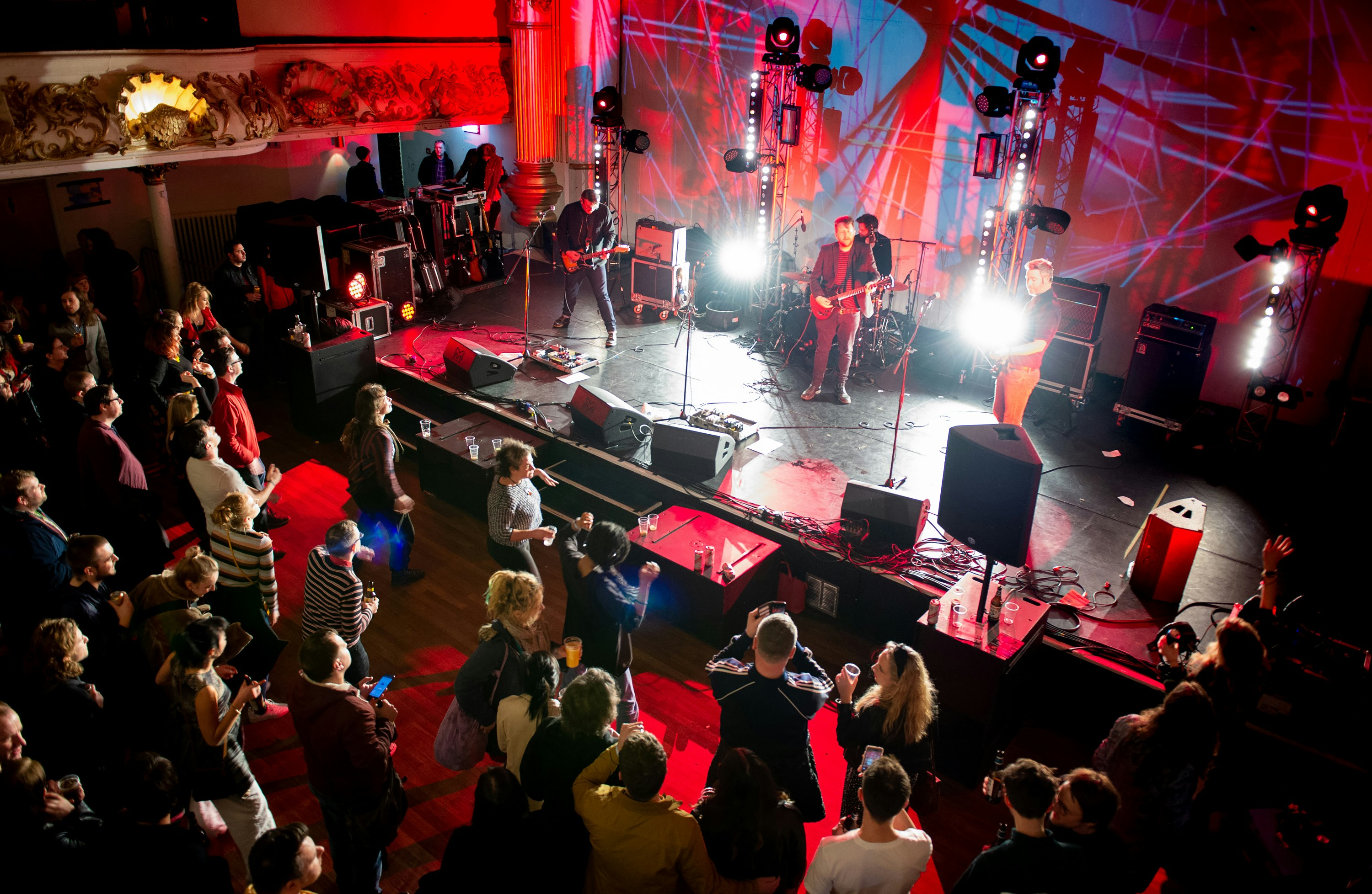 A three-piece band rock band play to a crowd at an intimate indoor concert. The band are in the spotlight while the crowd are in shadow.