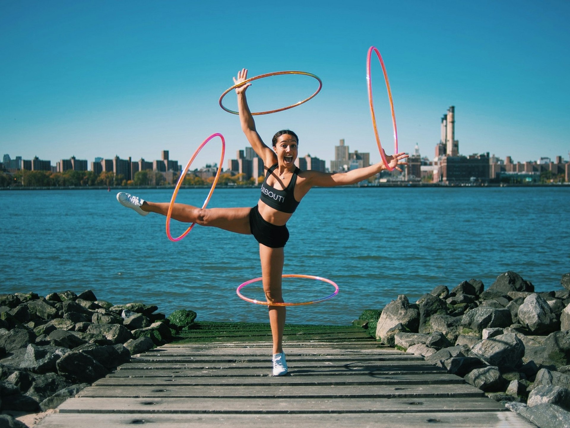 A woman with several hula hoops in motion on her arms, legs and body. She is smiling into the camera with water and a cityscape behind her.