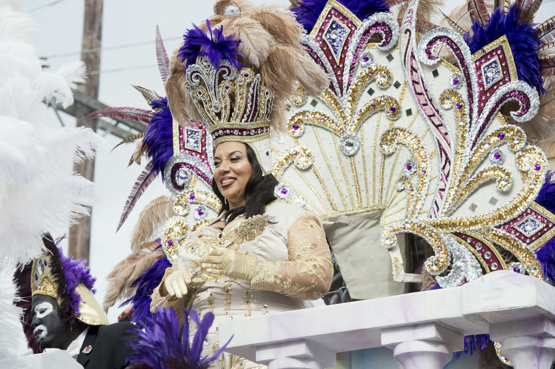 A woman dressed in an elaborately white and gold dress with sequined sleeves. Behind her is a large white, gold, blue and red sequined part of the float.