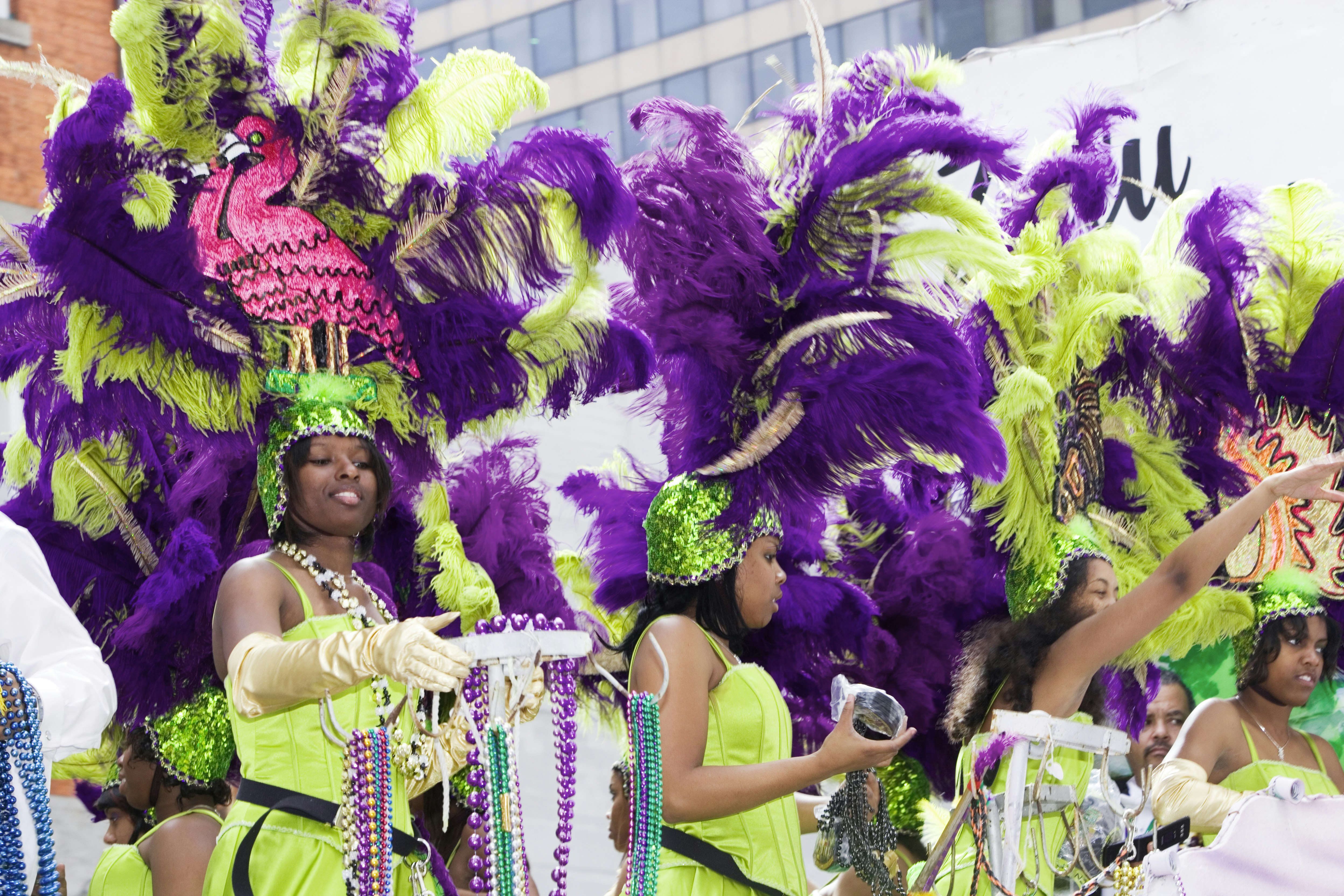 A group of women dressed in lime green dresses and green and purple feather and sequined headdresses throw beads to the crowd during Mardi Gras. 