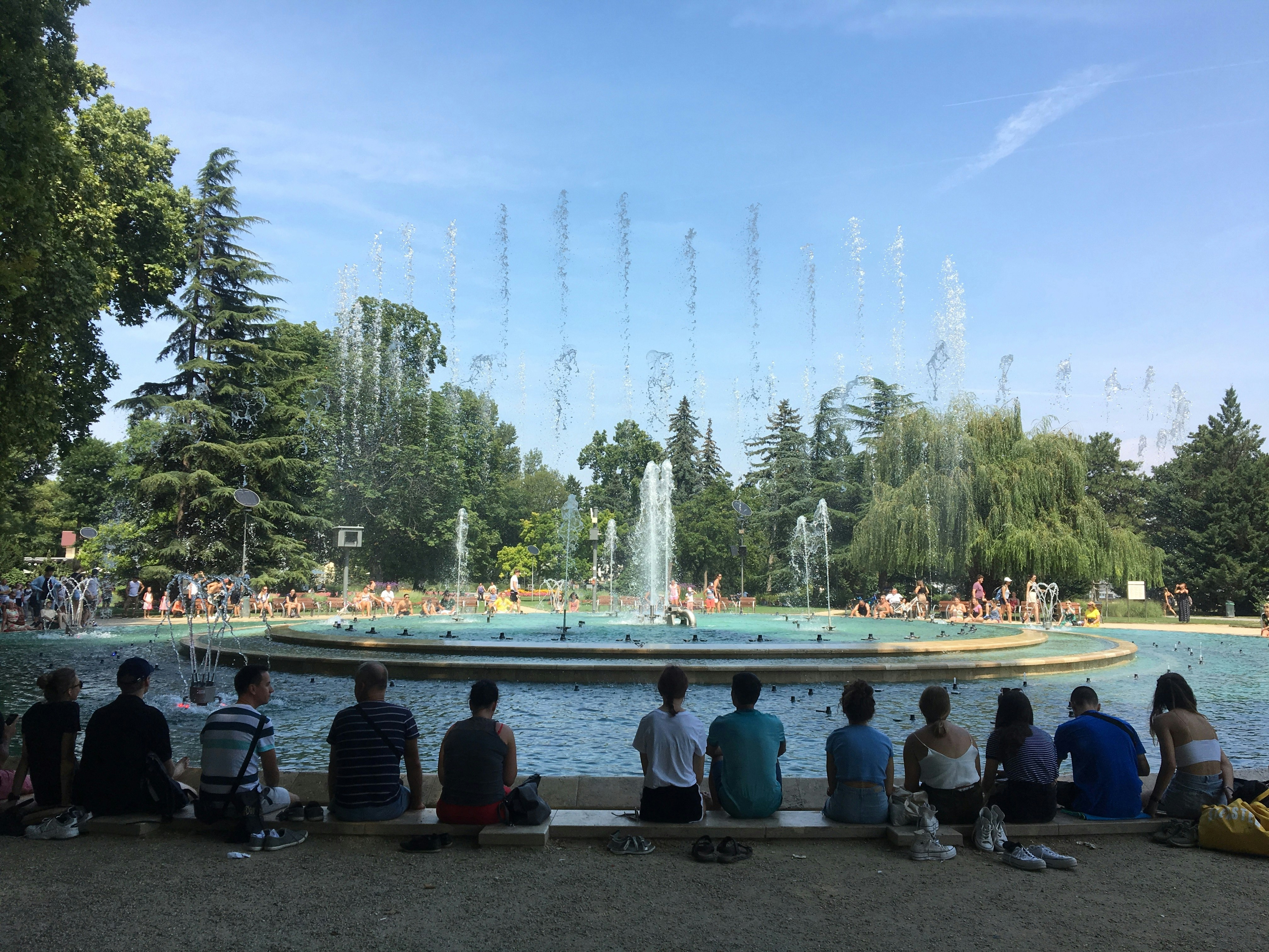 A row of people sit on he ground watching the fountain display on Margaret Island, Budapest. The fountain is shooting jets of water high into the air.