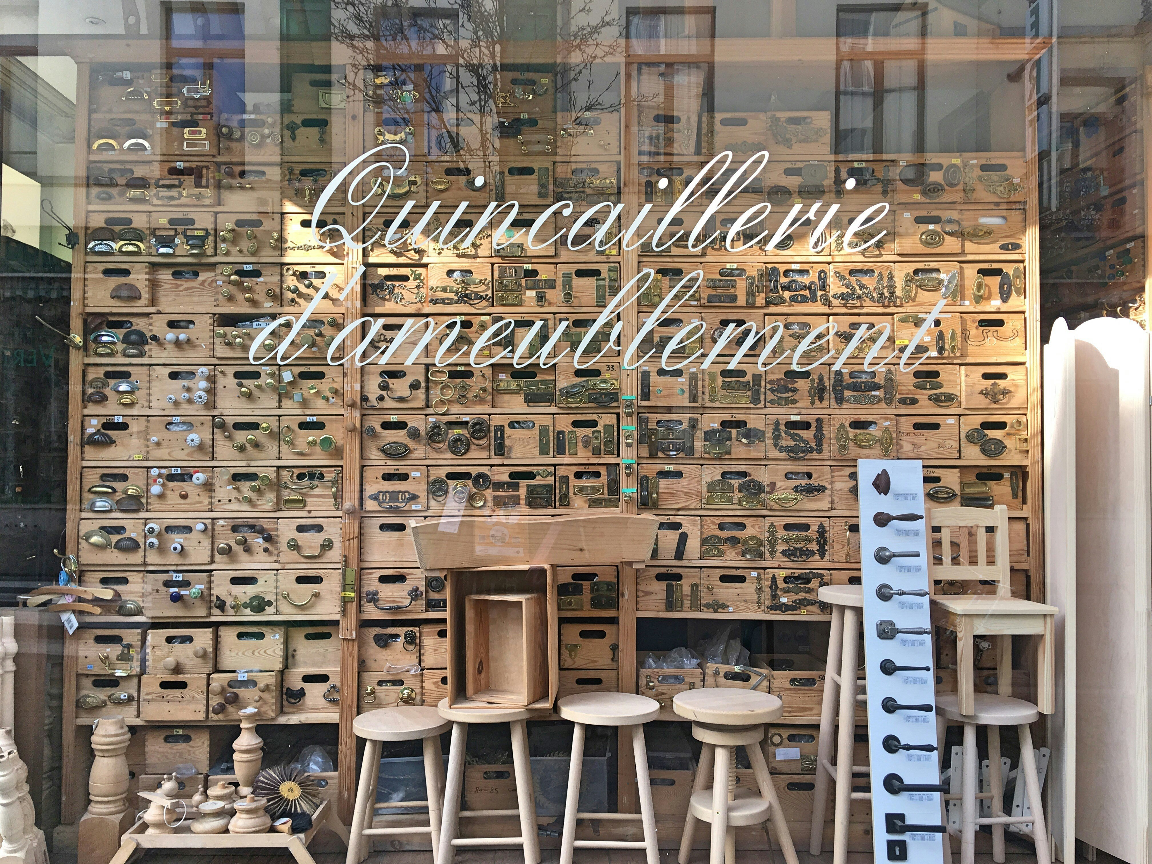 A shop window saying Quincaillerie d'ameublement in a cursive white script; the window contains rows and rows of wooden shelves adorned with handles, knobs and knockers; there are also stools and other wooden items on display.