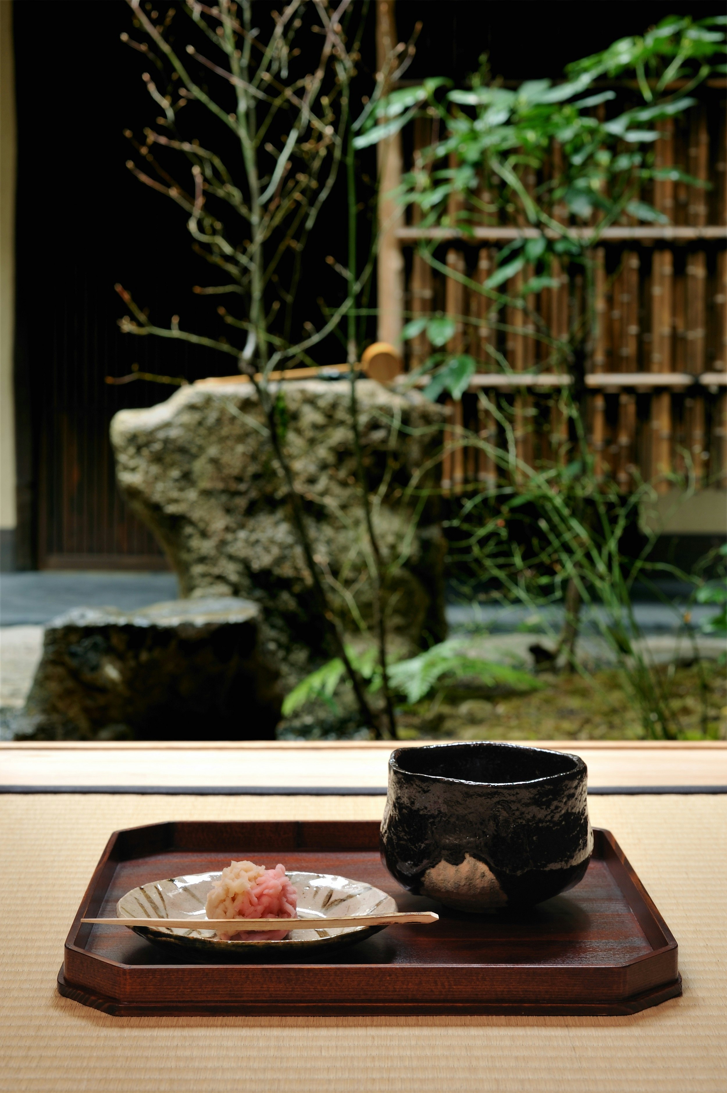 A bowl of matcha and wagashi sitting on a square wooden tray; these sit on a table in front of an open wall that reveals a rock-garden courtyard.