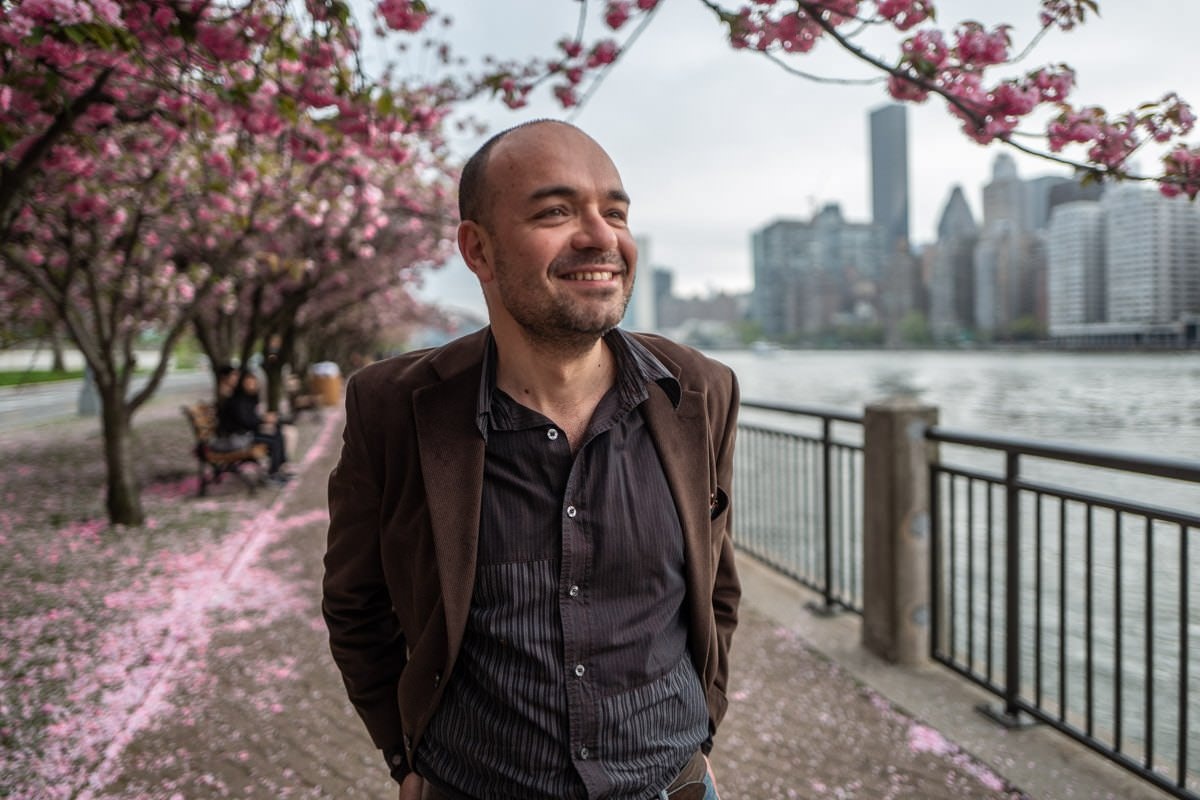 A photo of a man grinning, looking away from the camera. There is a river on the right and cherry blossom trees to the left with their petals swirling everywhere