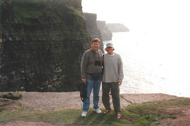 A father and son smile for the camera standing in front of the Cliffs of Moher in Ireland