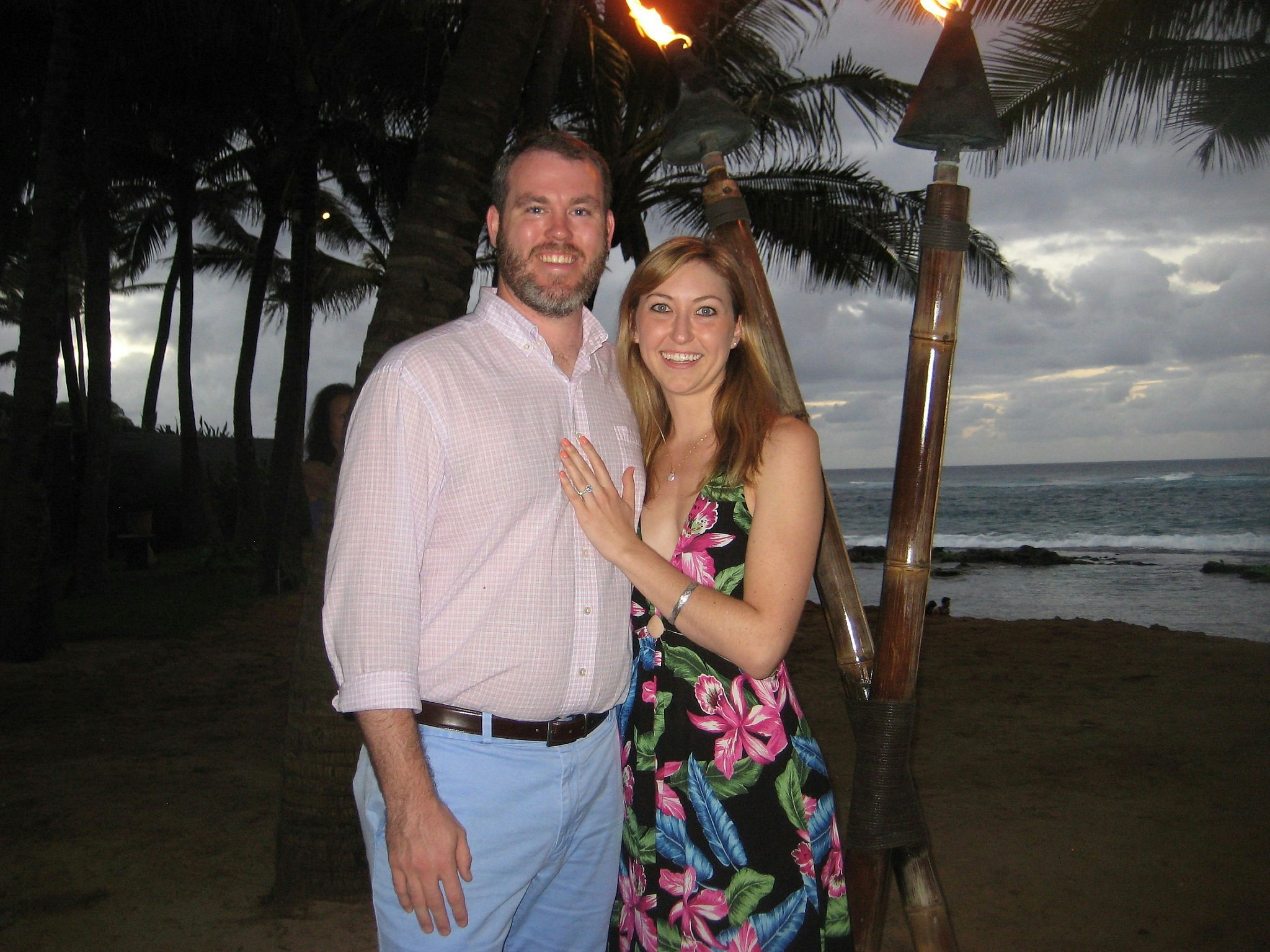Lonely Planet staffer Nicole and her husband stand on a beach in Maui at dusk with palm trees and the ocean behind them; the couple look happy and Nicole rests her hand on her partner's chest to show off her engagement ring.