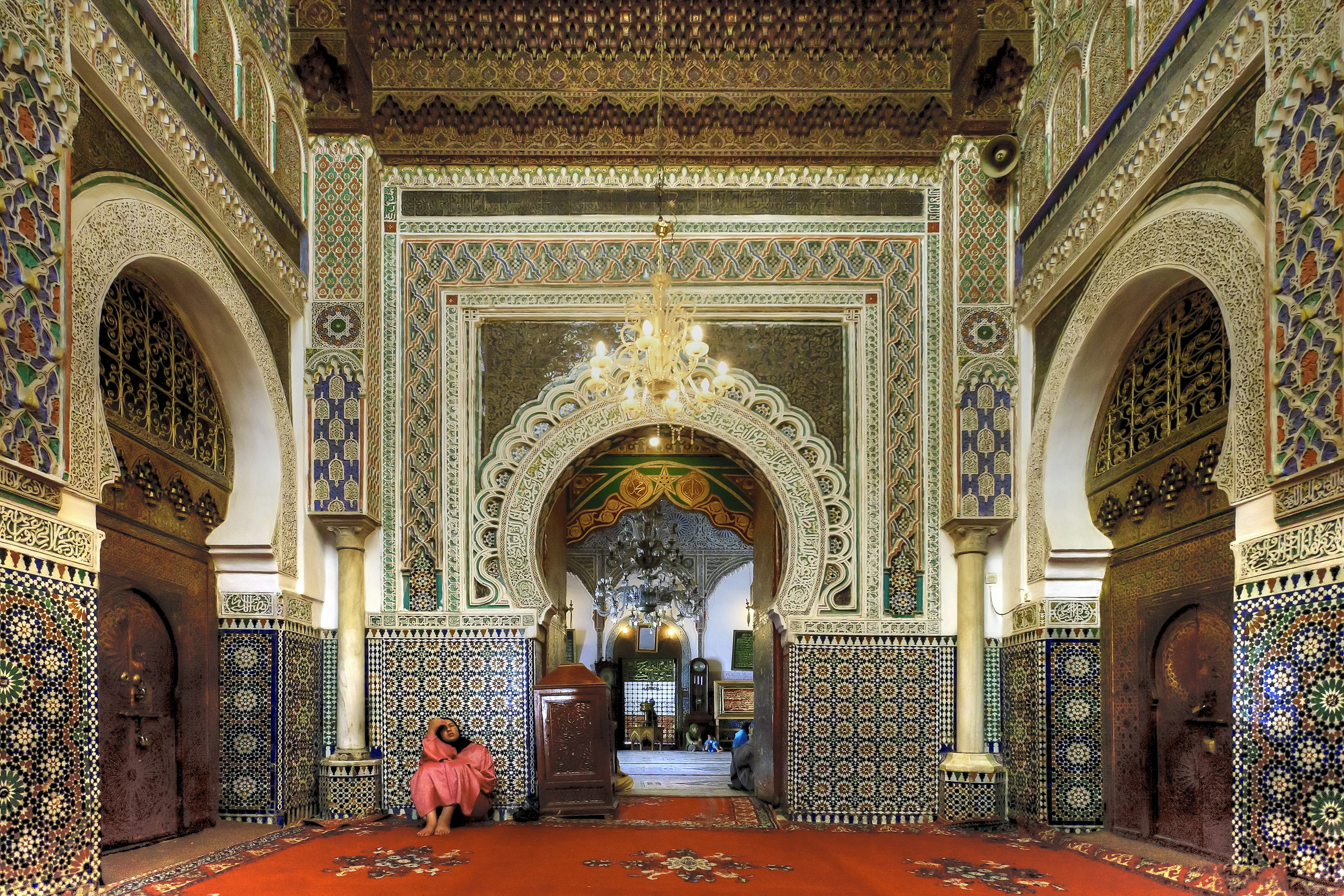 A woman in a long pink garment and black headscarf sits on the red-carpeted floor, touching her forehead, surrounded by the intricate mosaics and round, arched doors in the elaborate Mausoleum of Moulay 