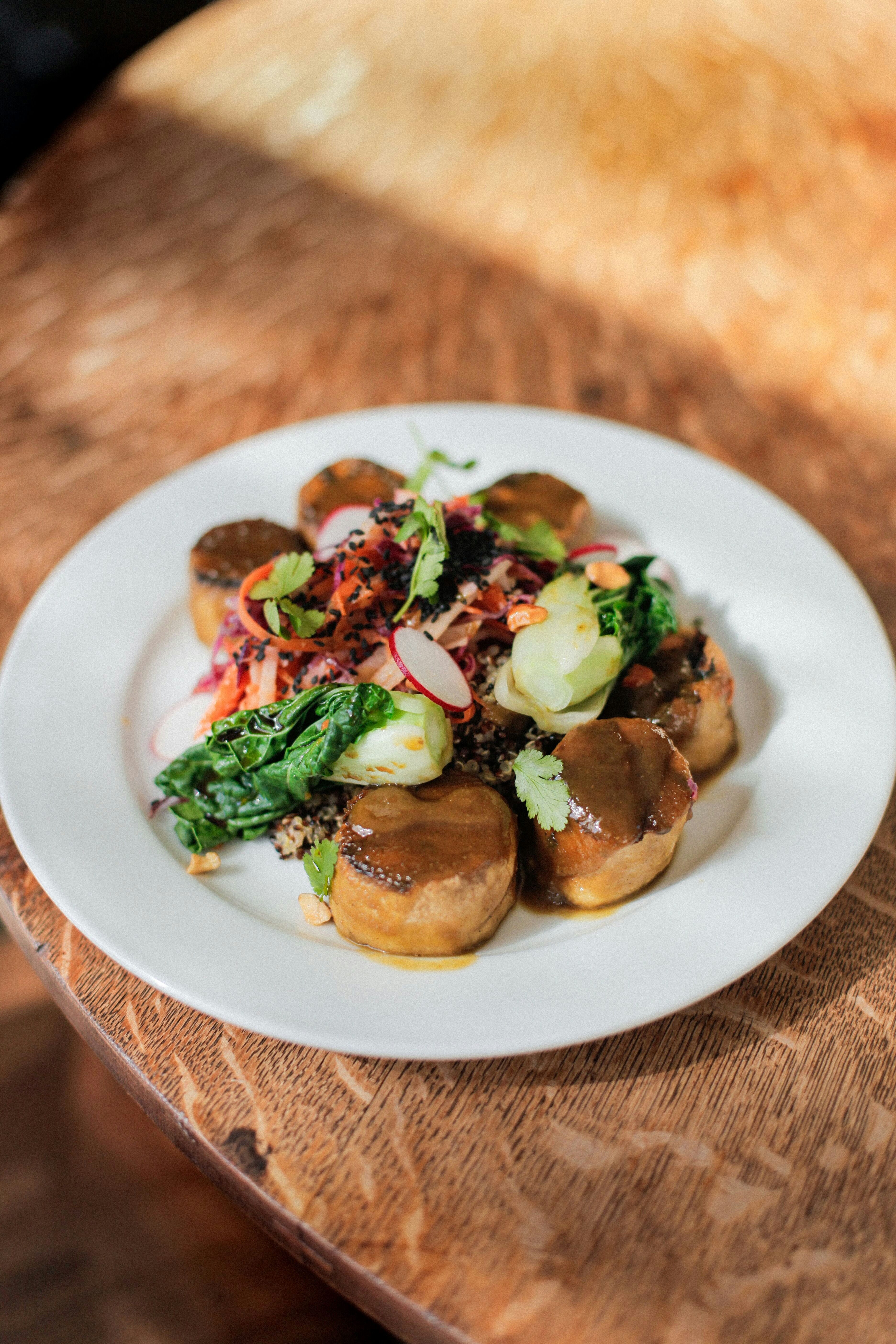 A plate of medallion-cut plantains are topped with a dark-colored sauce, shredded carrots, radishes, bok choy and green herbs. 