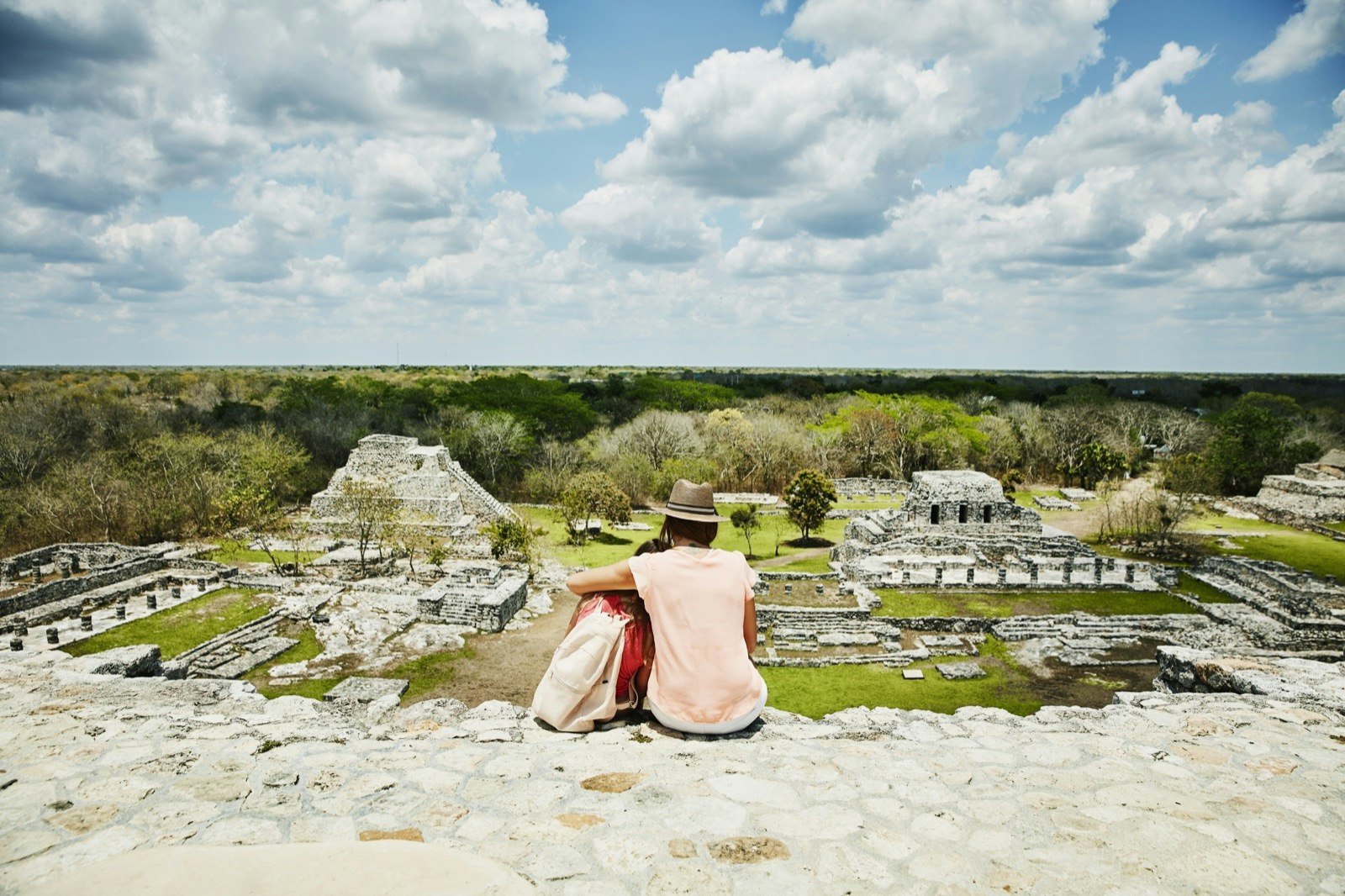 A woman and young girl embrace while sitting on a stone surface and looking out over the ruins of Mayapan 