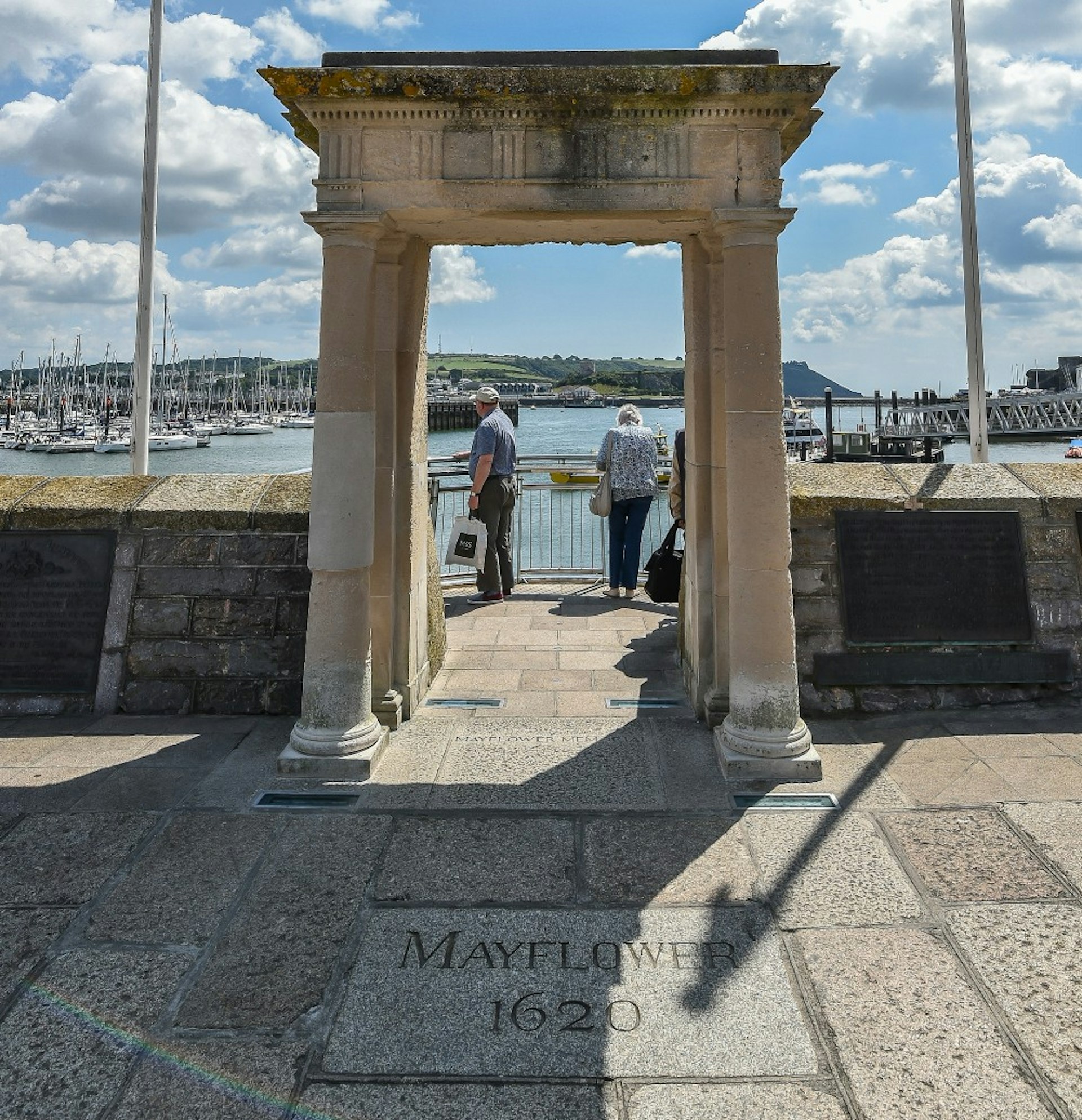 Two people look out over Plymouth harbour from the The Mayflower Steps on a sunny day.