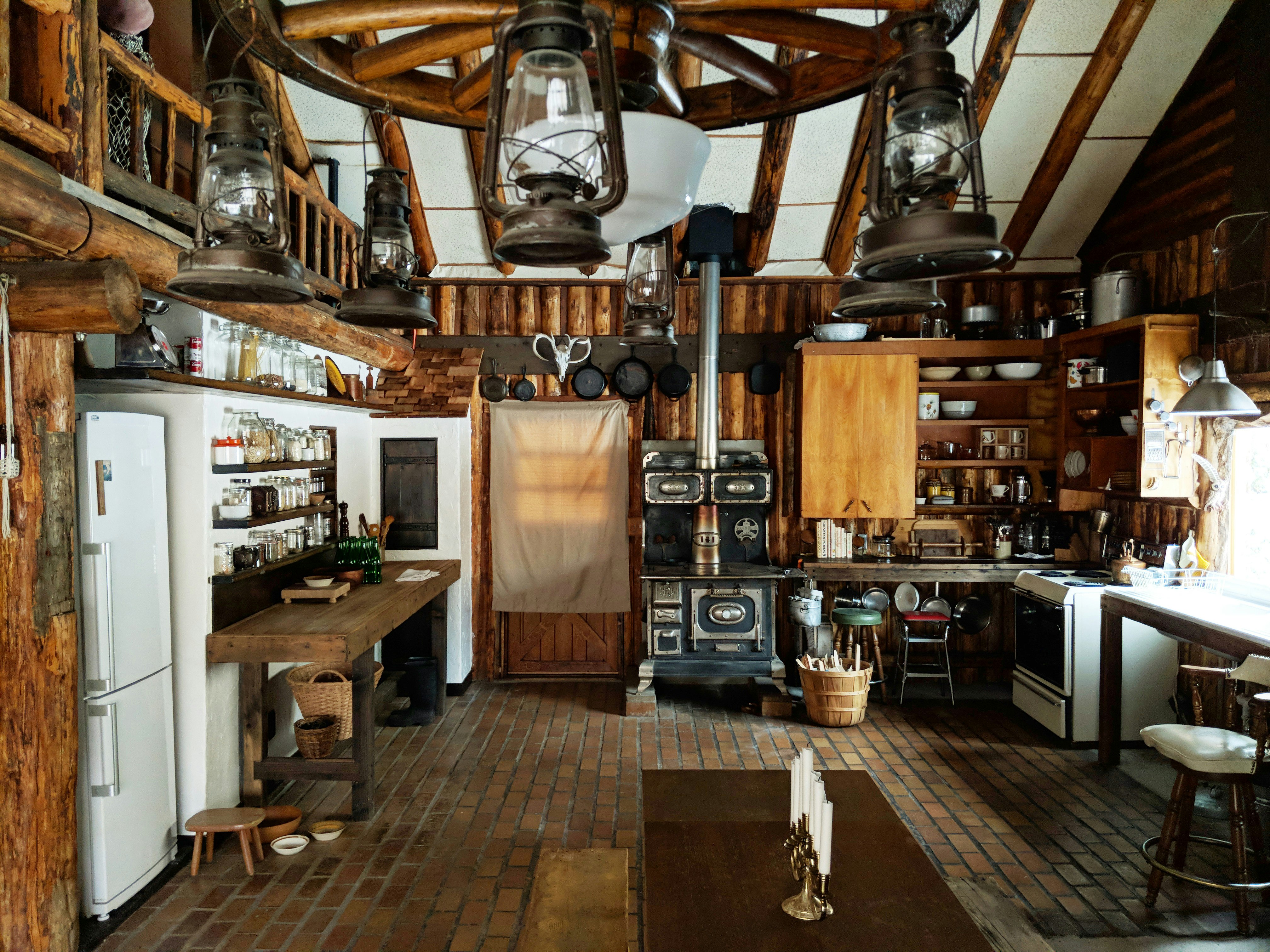 The large, open kitchen area of a cabin. The room is artistically cluttered, with a mixture of mis-matched furniture, and open shelving display crockery and mason jars filled with ingredients.
