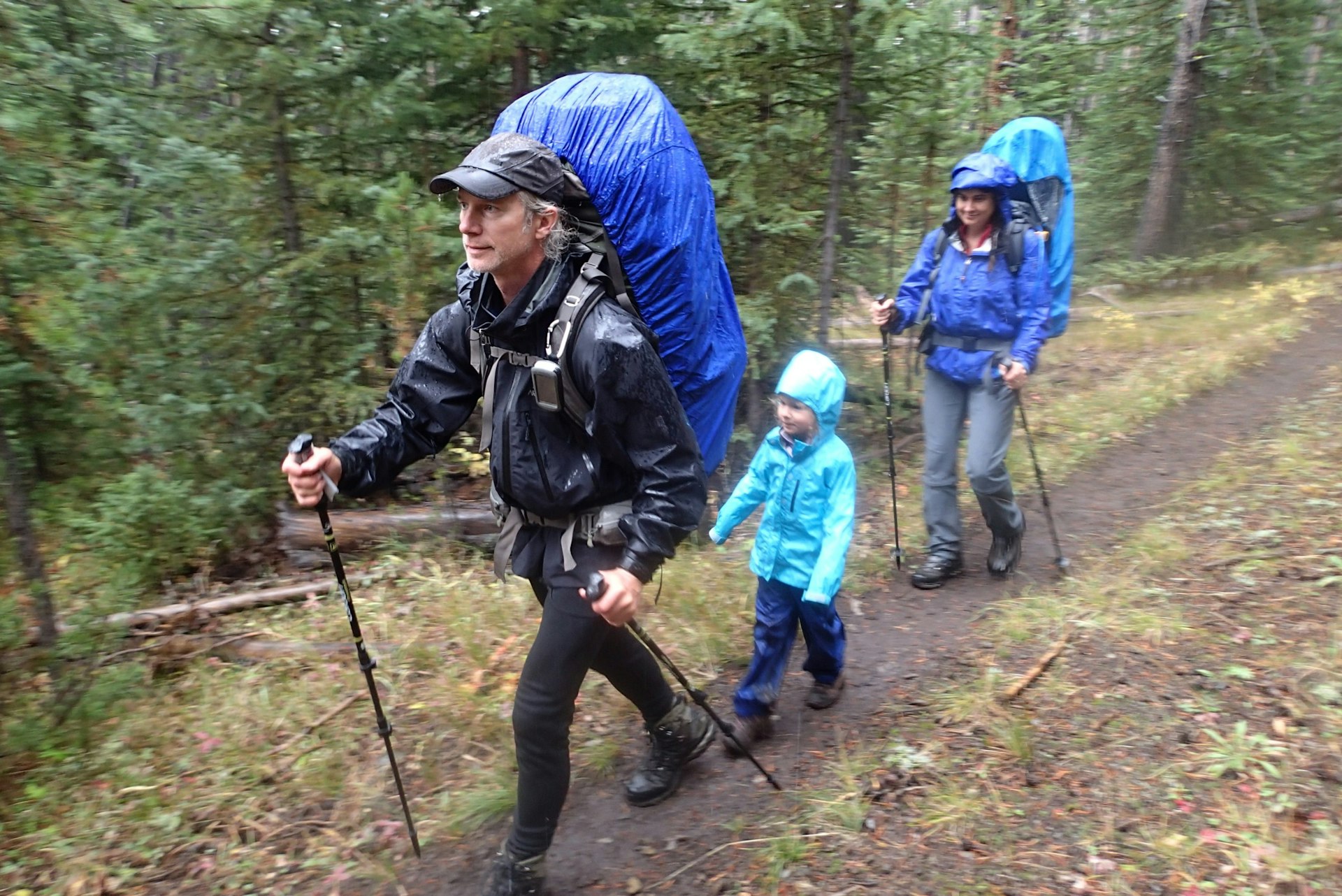 A man a woman and their daughter hike through a drizzle with backpacks and rain gear; Project Remote