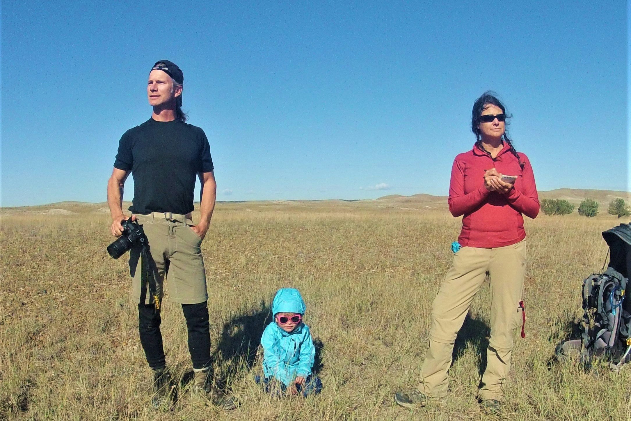 A man and a woman take photos and notes in a grassy plain as their daughter plays at their feet; Project Remote