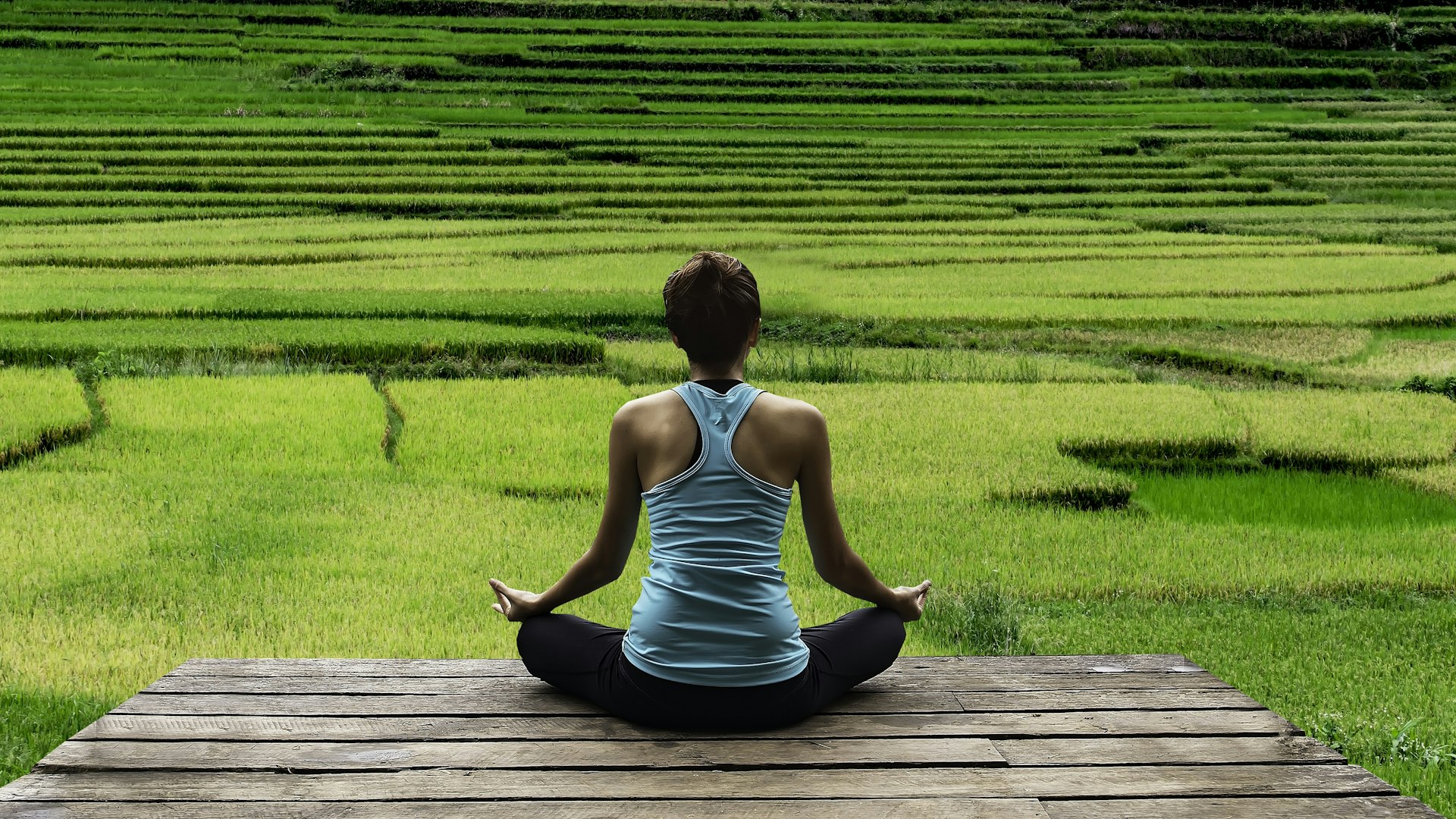 A woman, with her back to camera, sits in a meditative pose with her legs crossed in front of green rice fields in Bali.