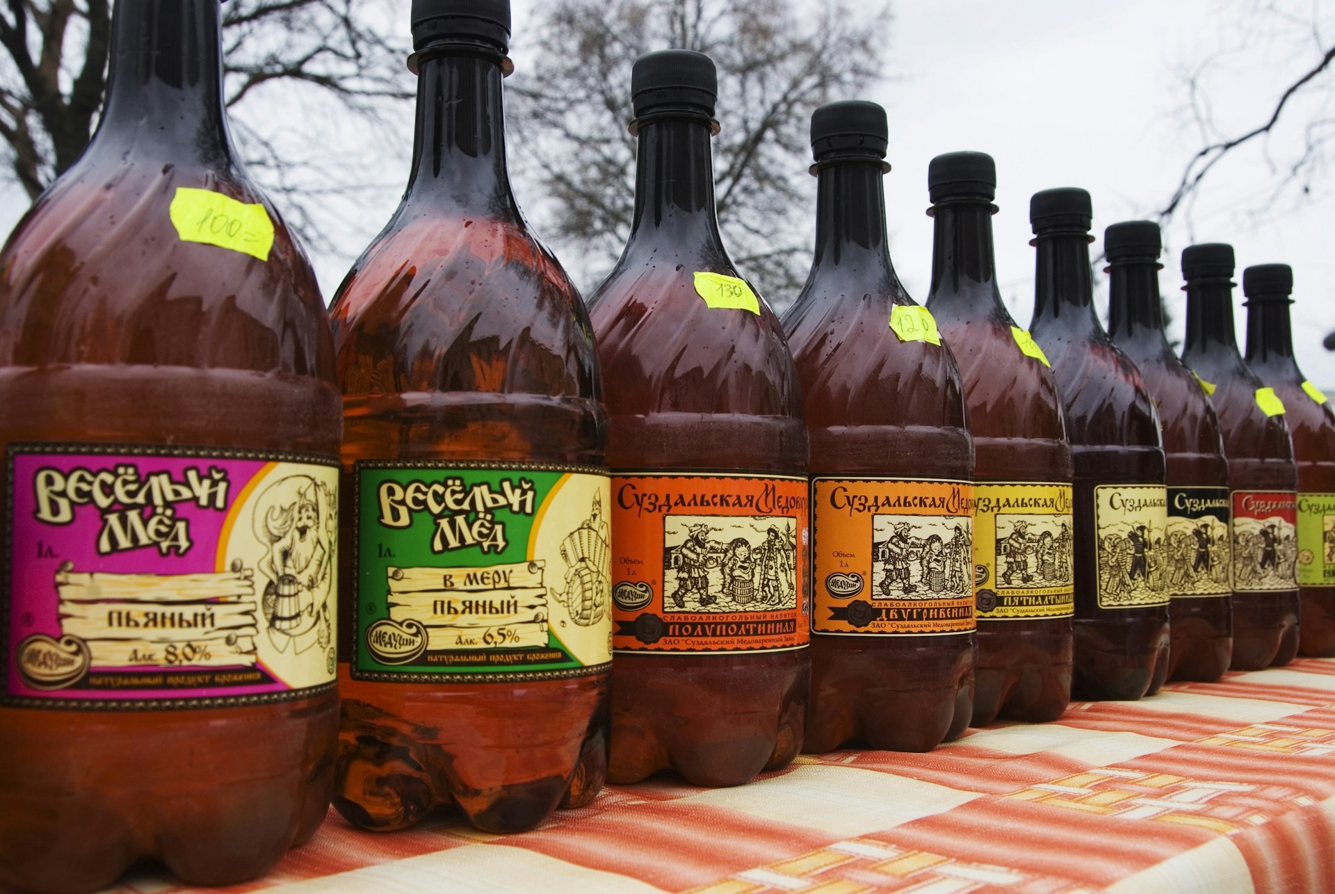 A row of brown bottles with colourful labels and Russian writing, sit on a red-and-white checked table cloth.