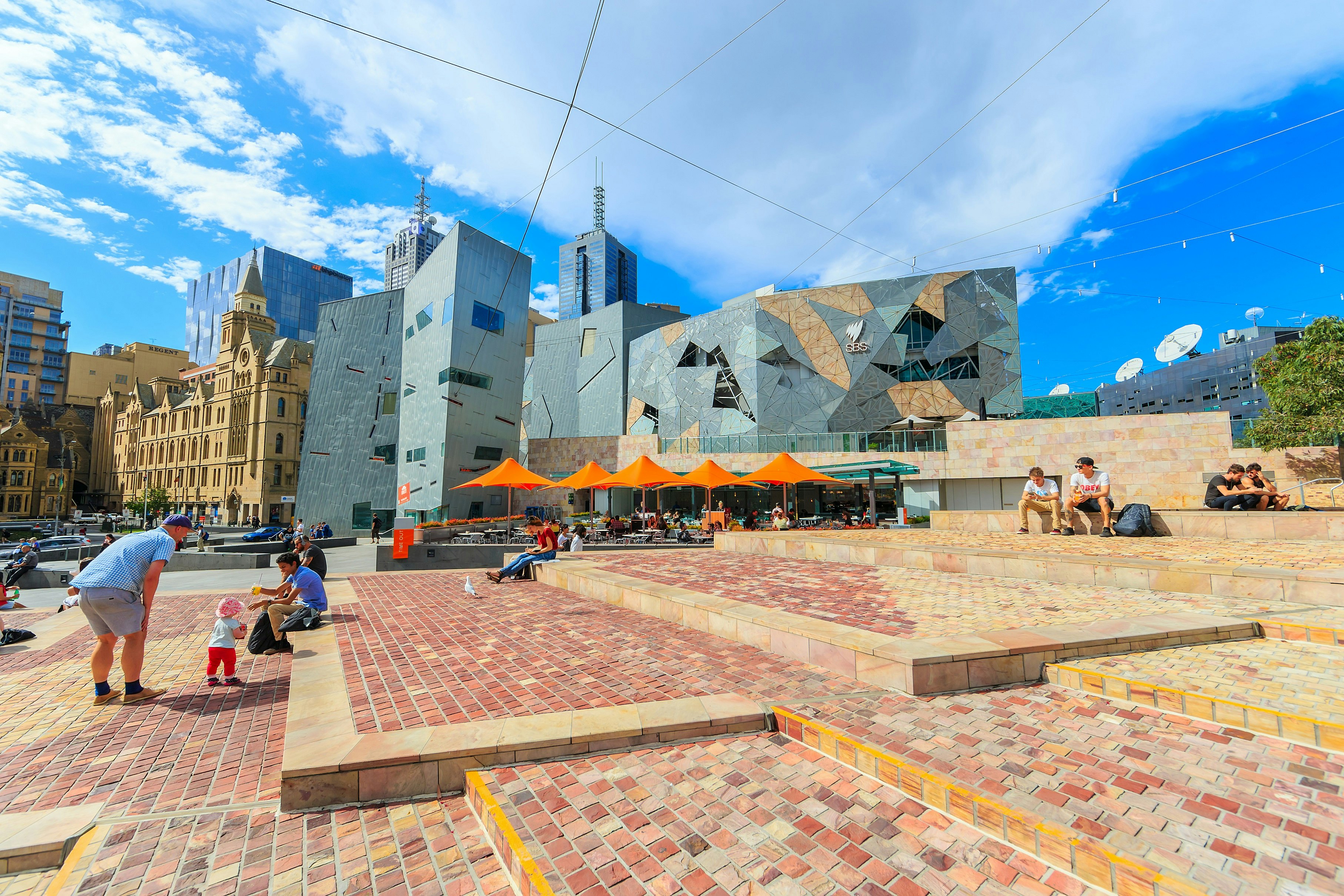 Federation Square in Melbourne, a mixed-use development in the inner city. Funky-looking buildings ring the square while many people converge on a large concrete structure in the centre, with space for sitting.