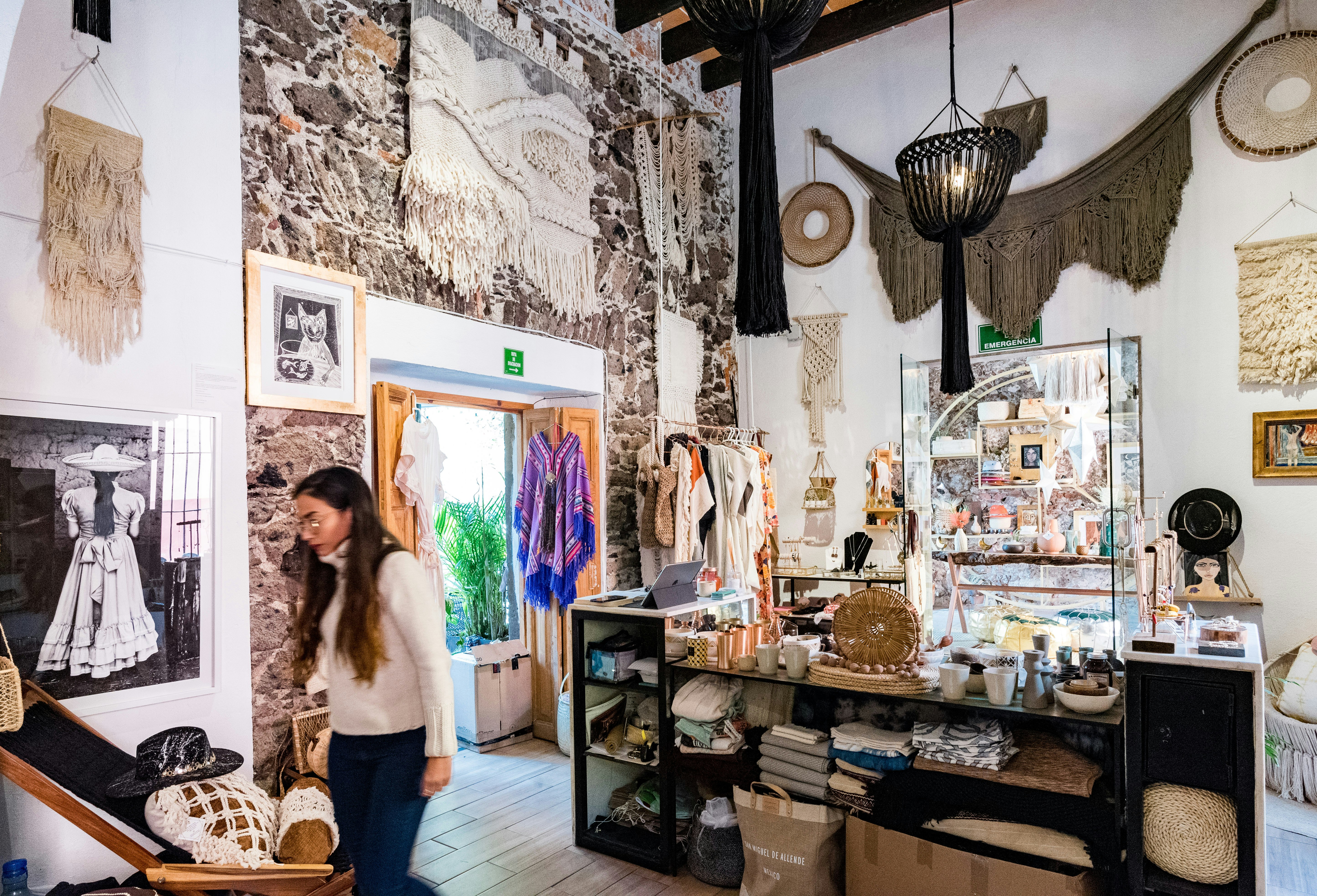 A woman with long brown hair, wearing a cream-colored sweater and jeans, walks through the interior of Mercado Collective, an interior design shop whose stone and white-washed walls are covered in hanging fabrics, with racks of clothing and jewelry visible in the background
