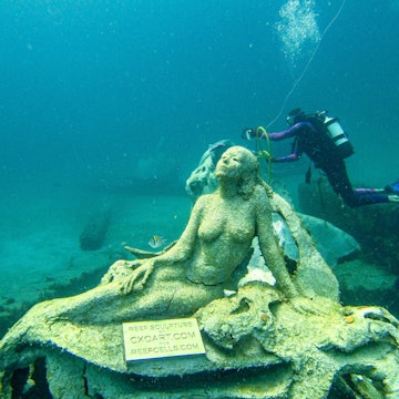 Underwater shot of a mermaid sculpture at the bottom of the ocean in Florida. In the background, a scuba diver appears to measuring something.
