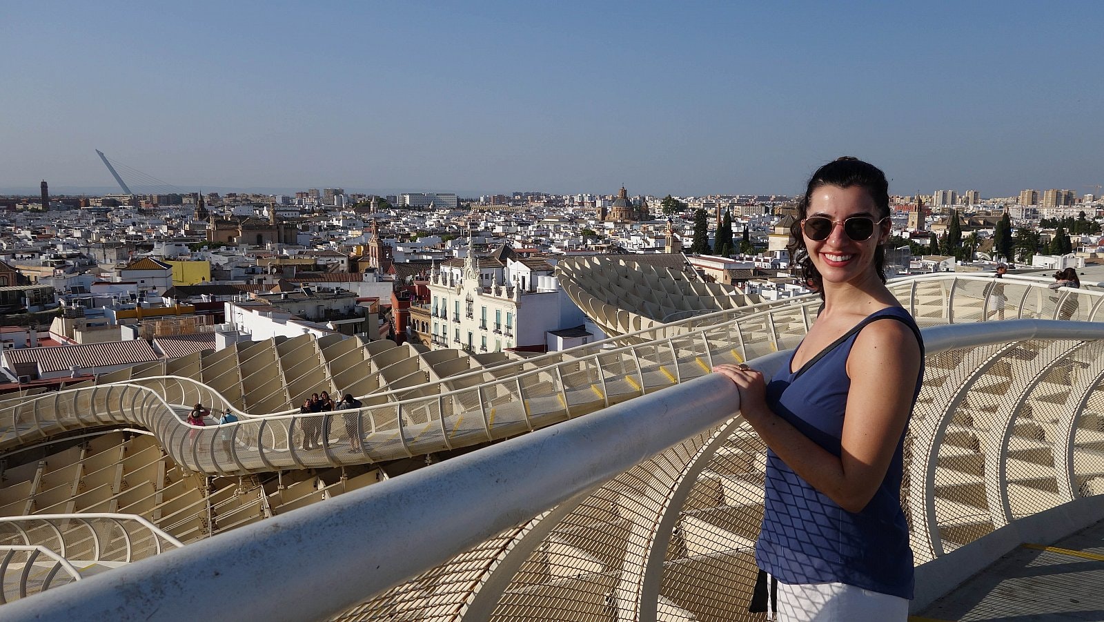 Writer Caterina stands on the undulating wooden walkway atop the Metropol Parasol, wearing sunglasses and smiling with the Seville skyline behind her.