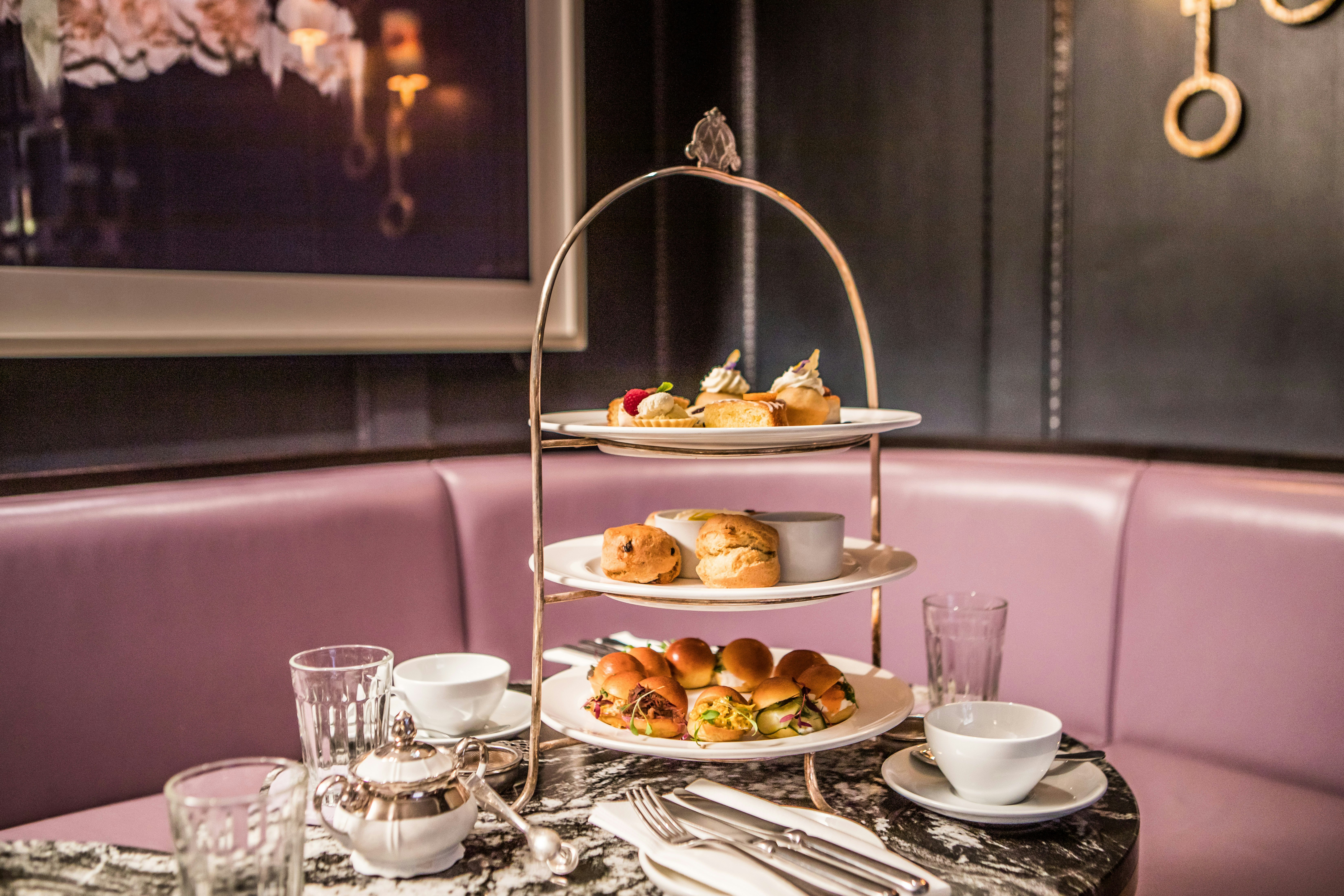 A platter of cakes and sandwiches on a marbled table, with pink corner seating in the background.