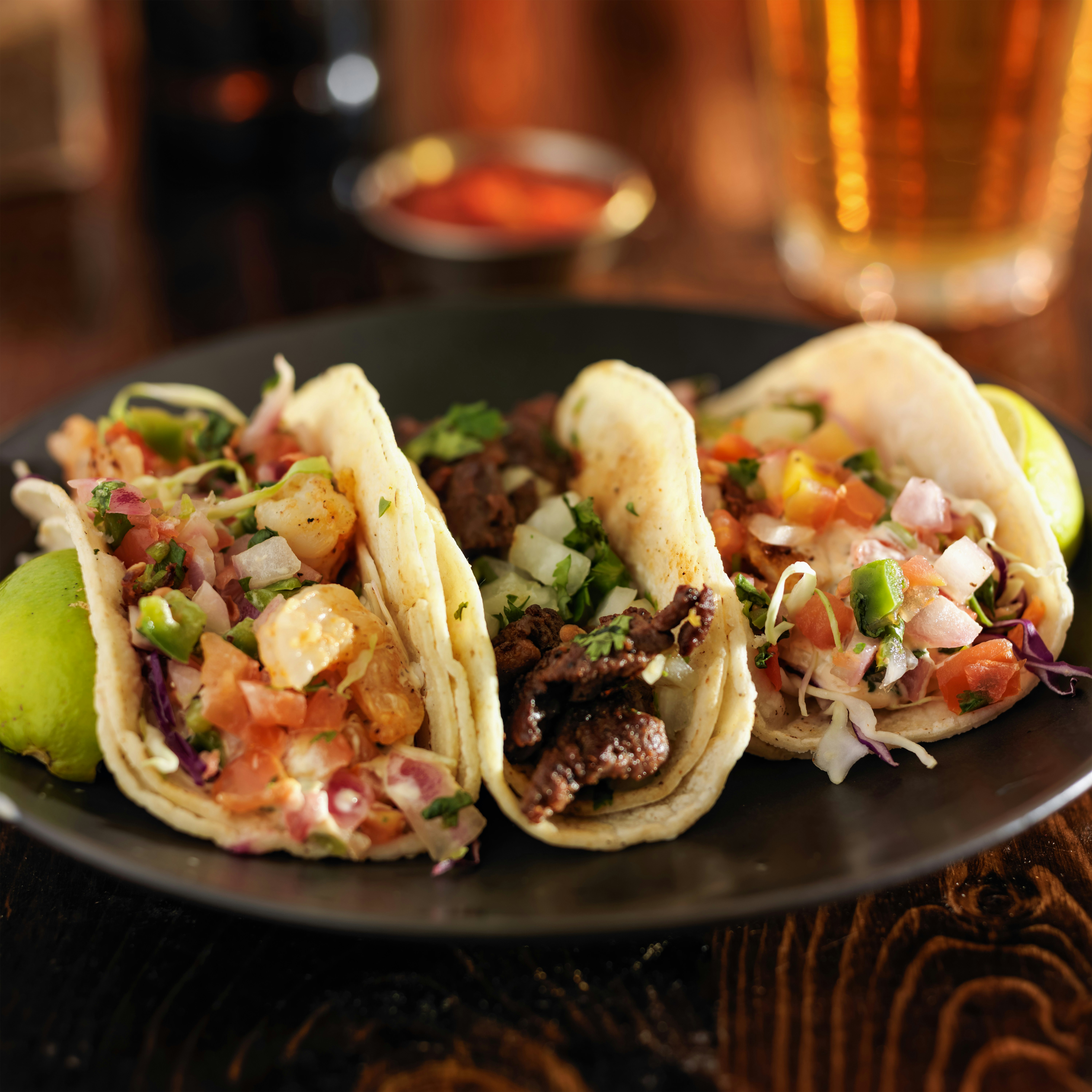 A trio of soft-shell tacos each individually filled with shrimp, steak and fish and topped with onions, cilantro and tomatoes. There is a pair of sliced limes on each side of the plate.