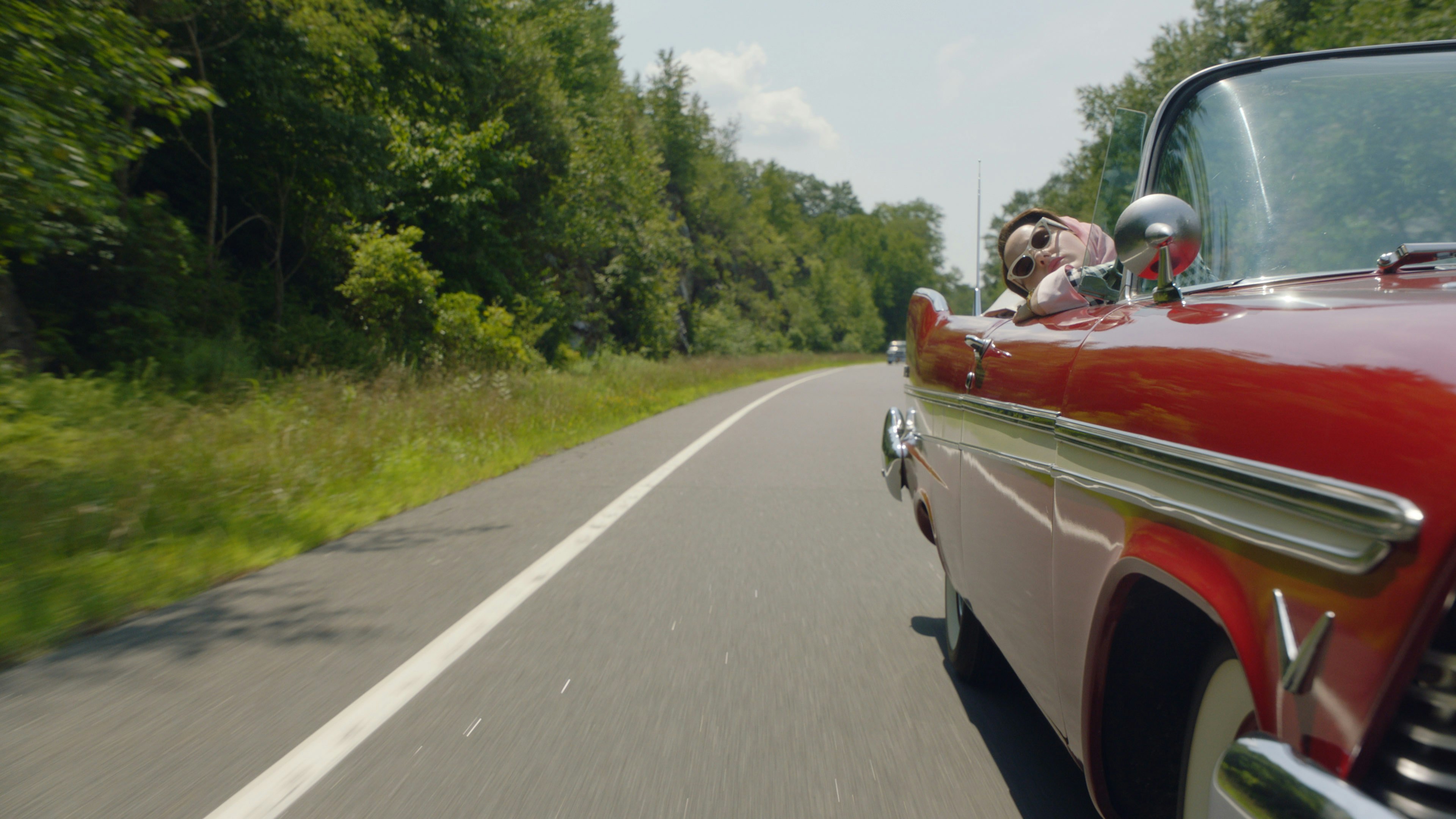 Midge Maisel in a classic red car driving through forests