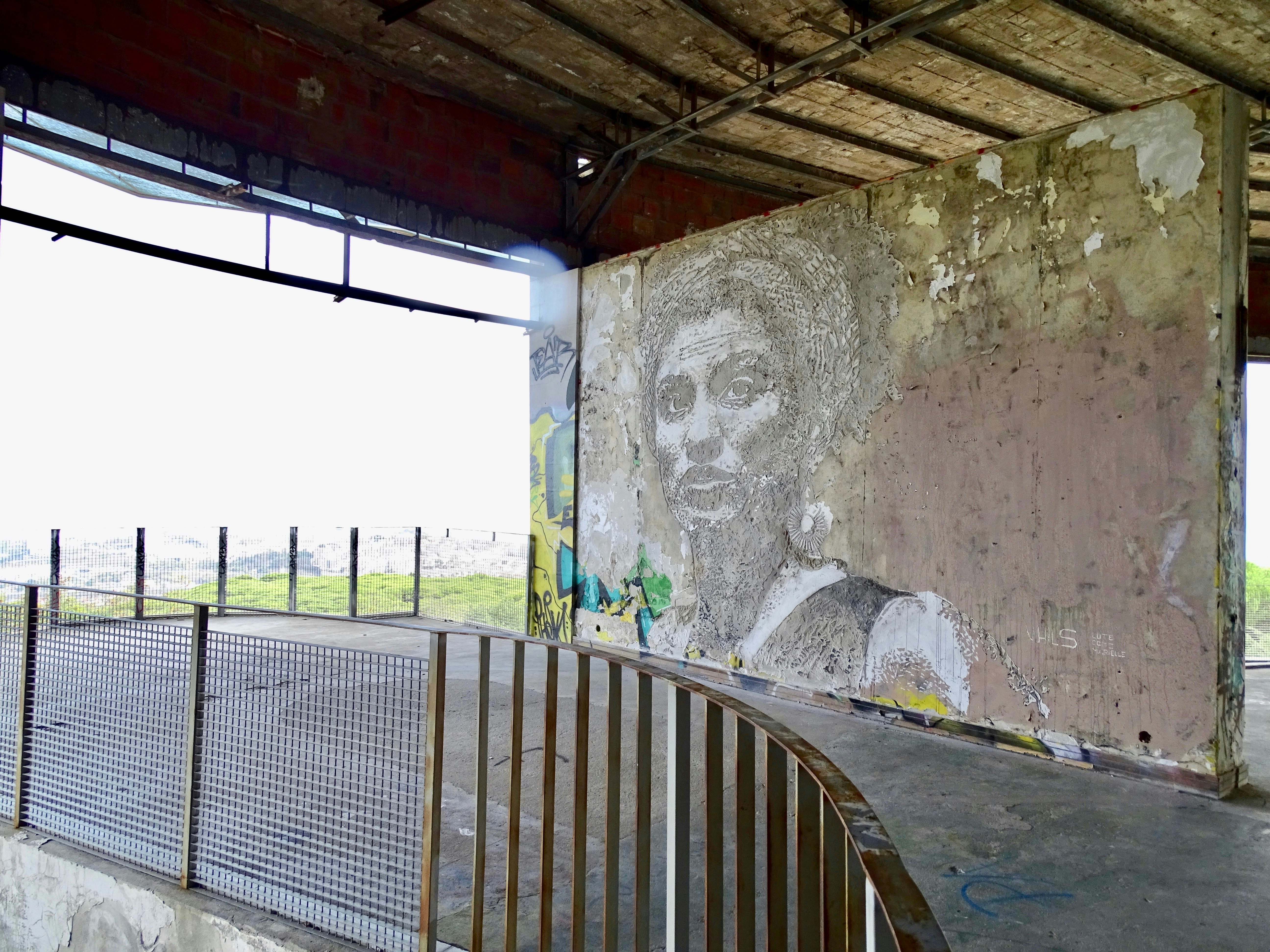 A black and white mural of a woman, taking up the whole cracked concrete wall with a view of the city behind her.
