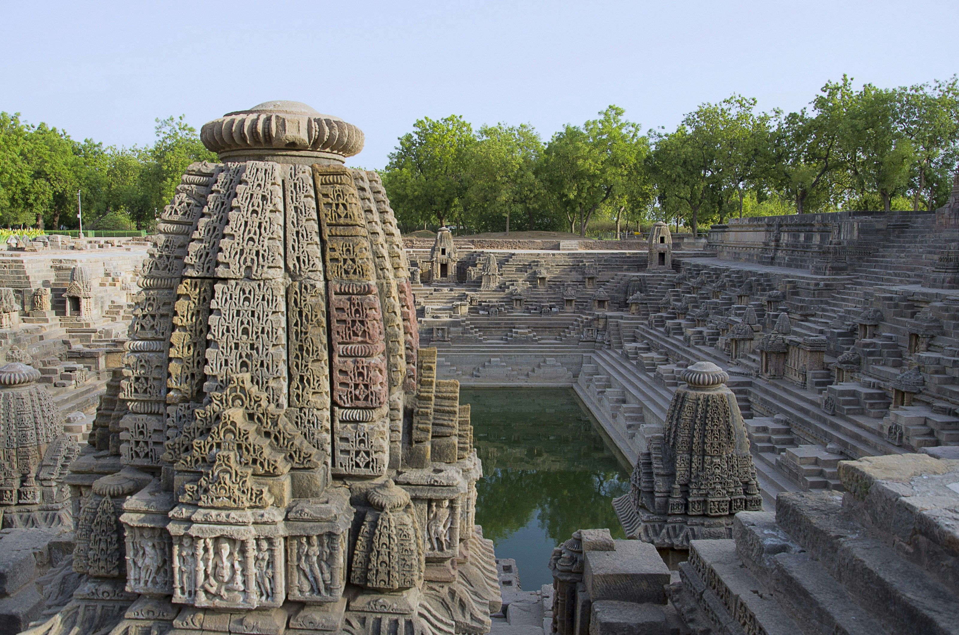 A view over the Surya Kund stepwell in Gujarat, with a close-up view of one of the small shrines dominating the image, and the large well, with the diagonal steps, visible behind it. In the background trees and other greenery are visible.