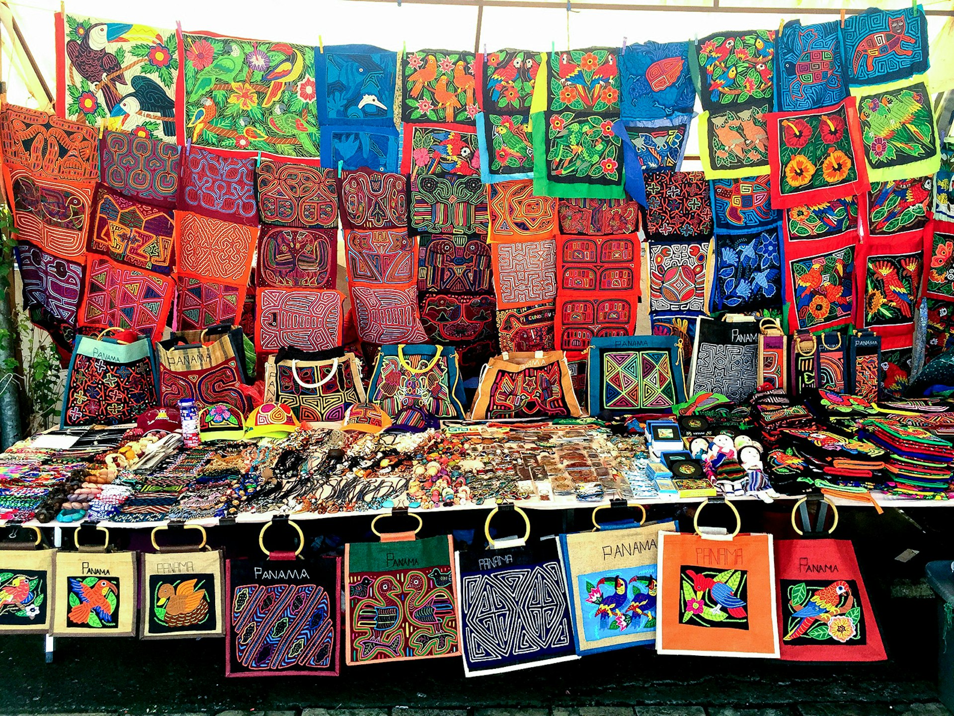 A stand filled with colorful and inA stand filled with colorful and intricately designed fabrics hang from a clothes hanger and are also laid out on a table. A collection of bags with wooden handles dangle from the front of the table.tricately designed fabrics hang from clothes hanger and are laid out on a table. A collection of bags with wooden handles dangle from the front of the table.