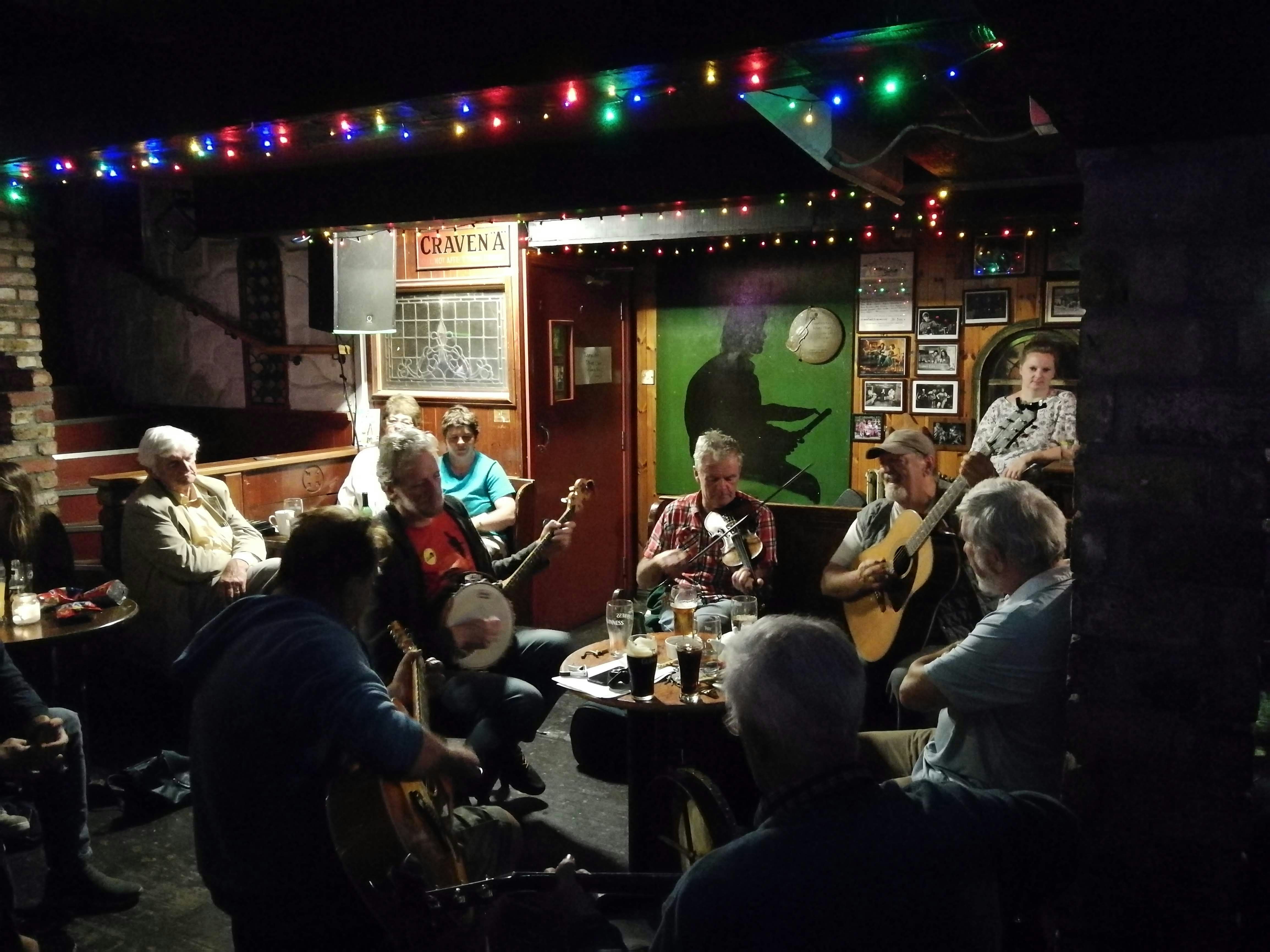 Inside a dimly lit pub, a group of folk musicians are playing around a table, as an audience looks on. Colourful fairy lights are hanging overhead.