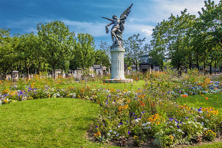 An angel statue stands above grave stones and a field of wildflowers
