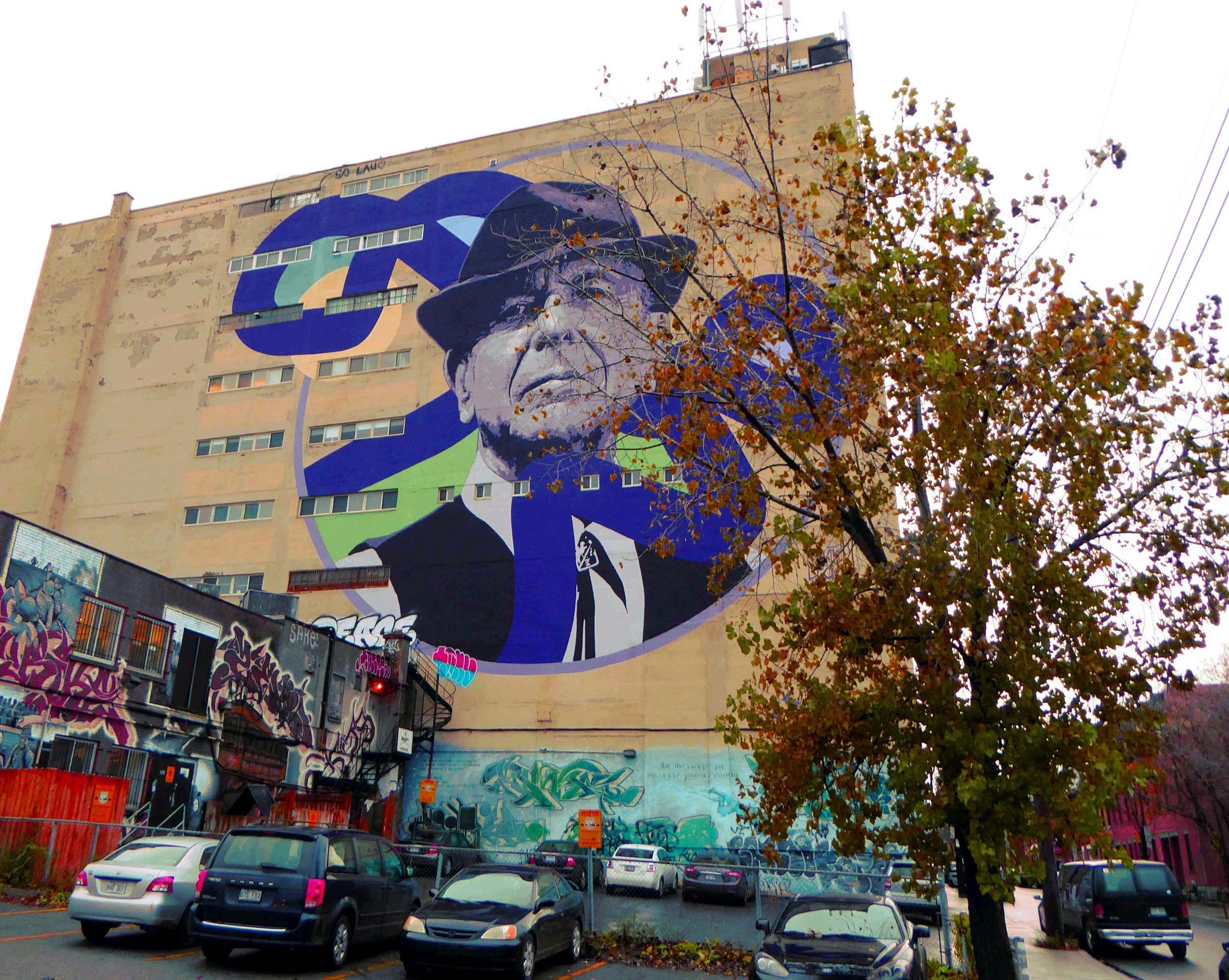 A large mural of Leonard Cohen is seen on the side of a building in Quebec. A tree partially obscuring the painting has autumn leaves turning brown; Montreal in the fall