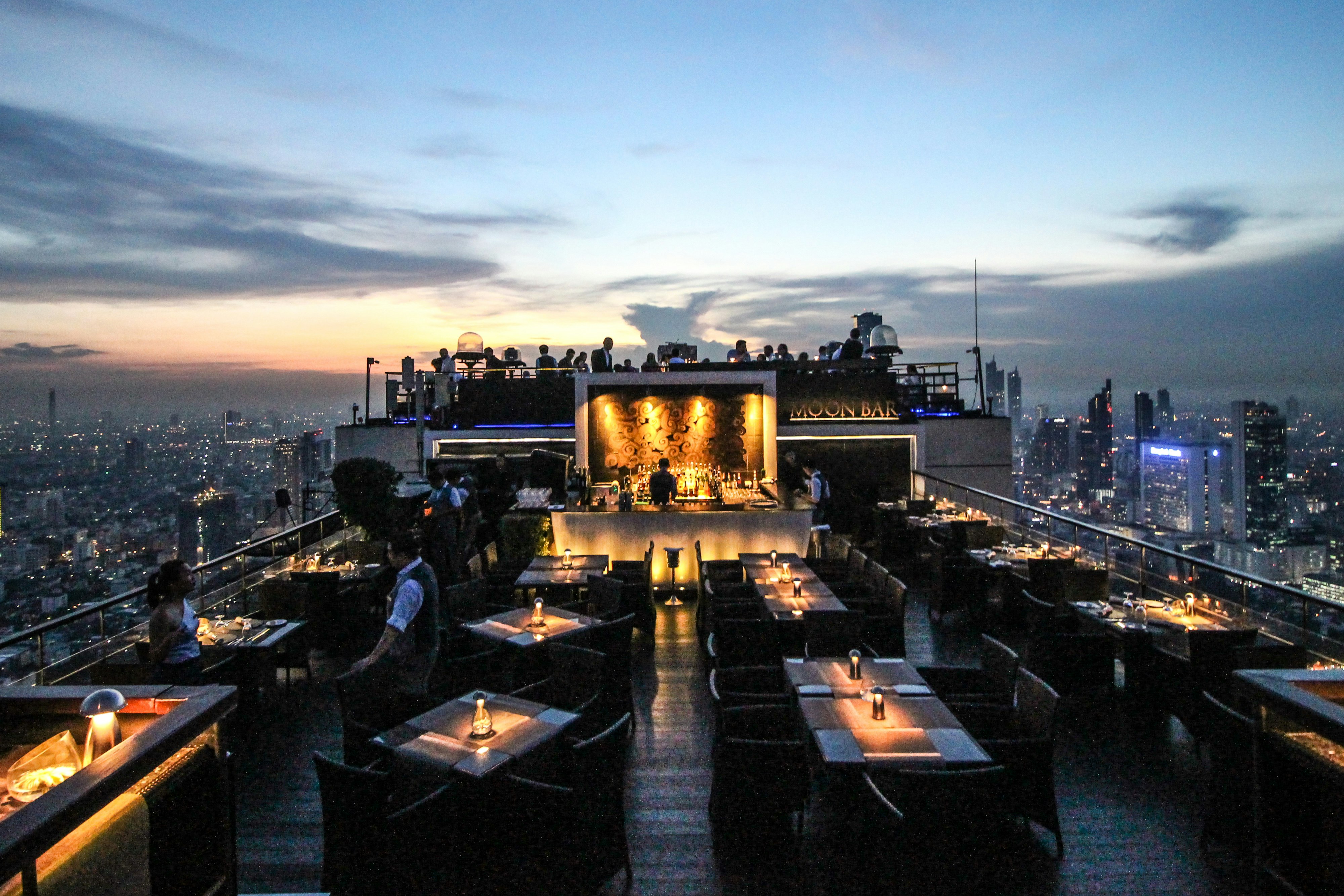 Patrons of Moon Bar are drinking and socialising at dusk. The Bangkok skyline is visible