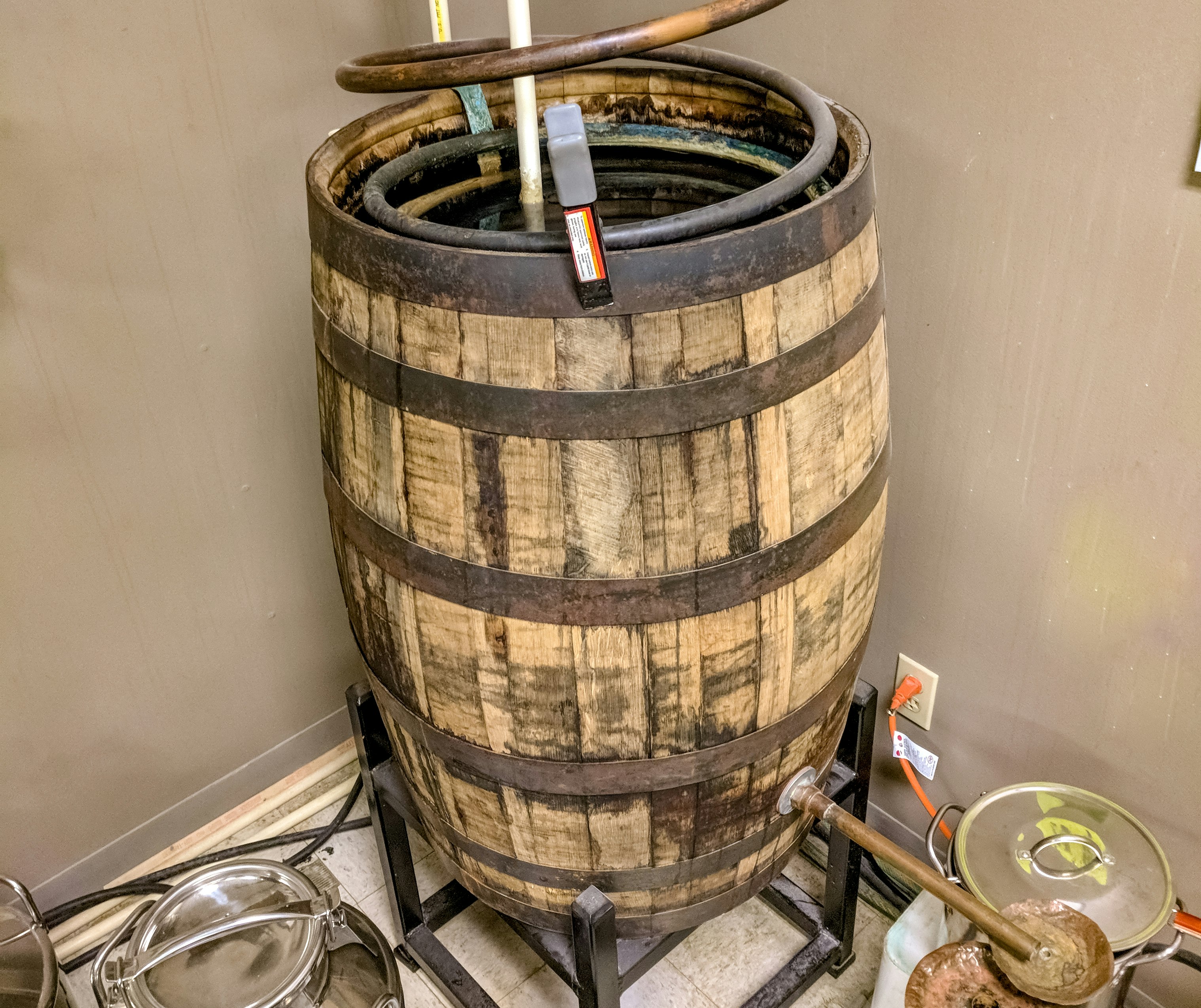 A large wooden barrel has a large wire copper coil inside of it. A long copper spout is attached to the bottom of the barrel. There are a collection of silver pots on the floor.