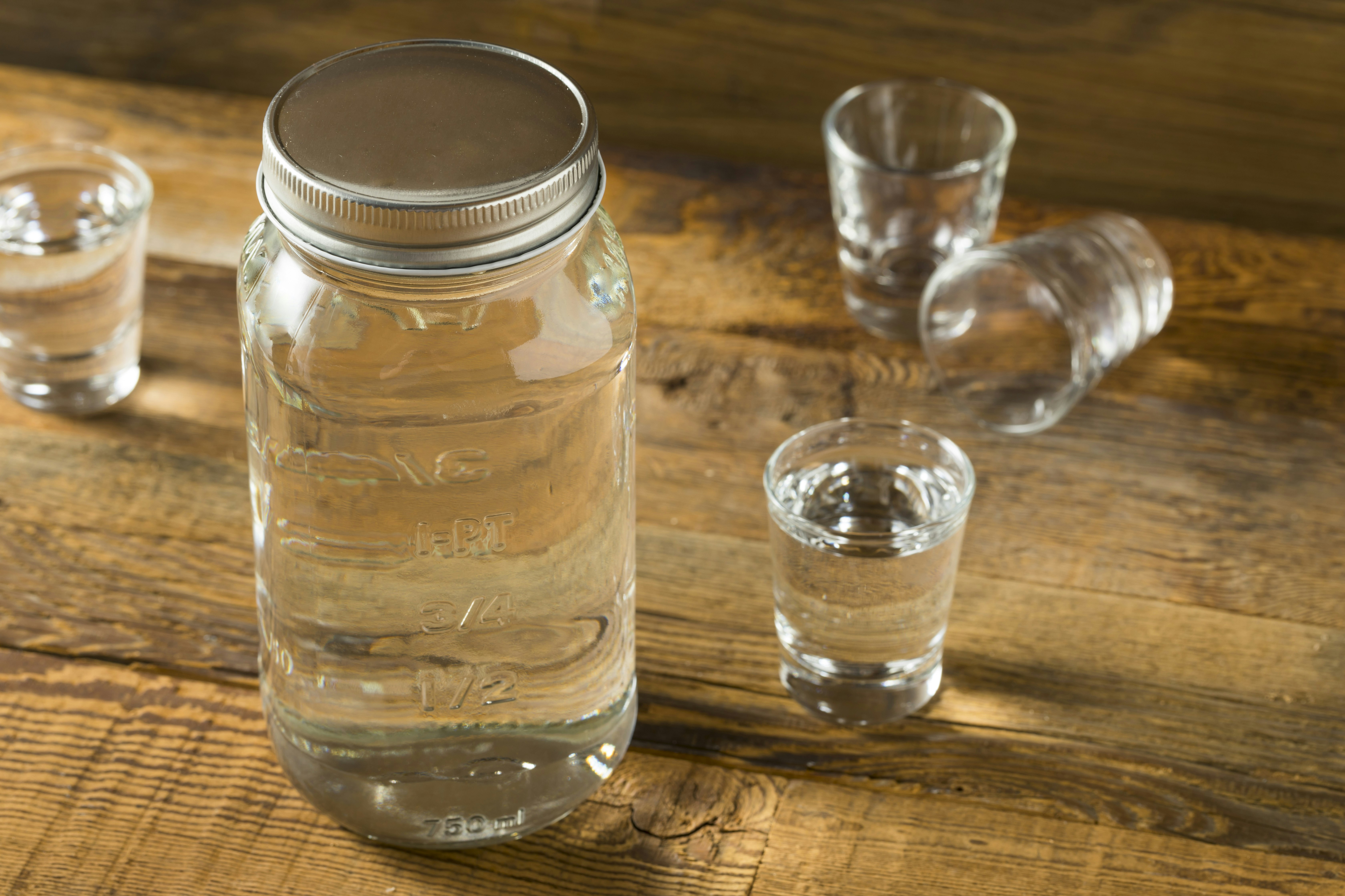A mason jar filled with a clear liquid. There are two shot glasses filled with the clear liquid. There is one empty shot glass and another empty shot glass lying on its side. 