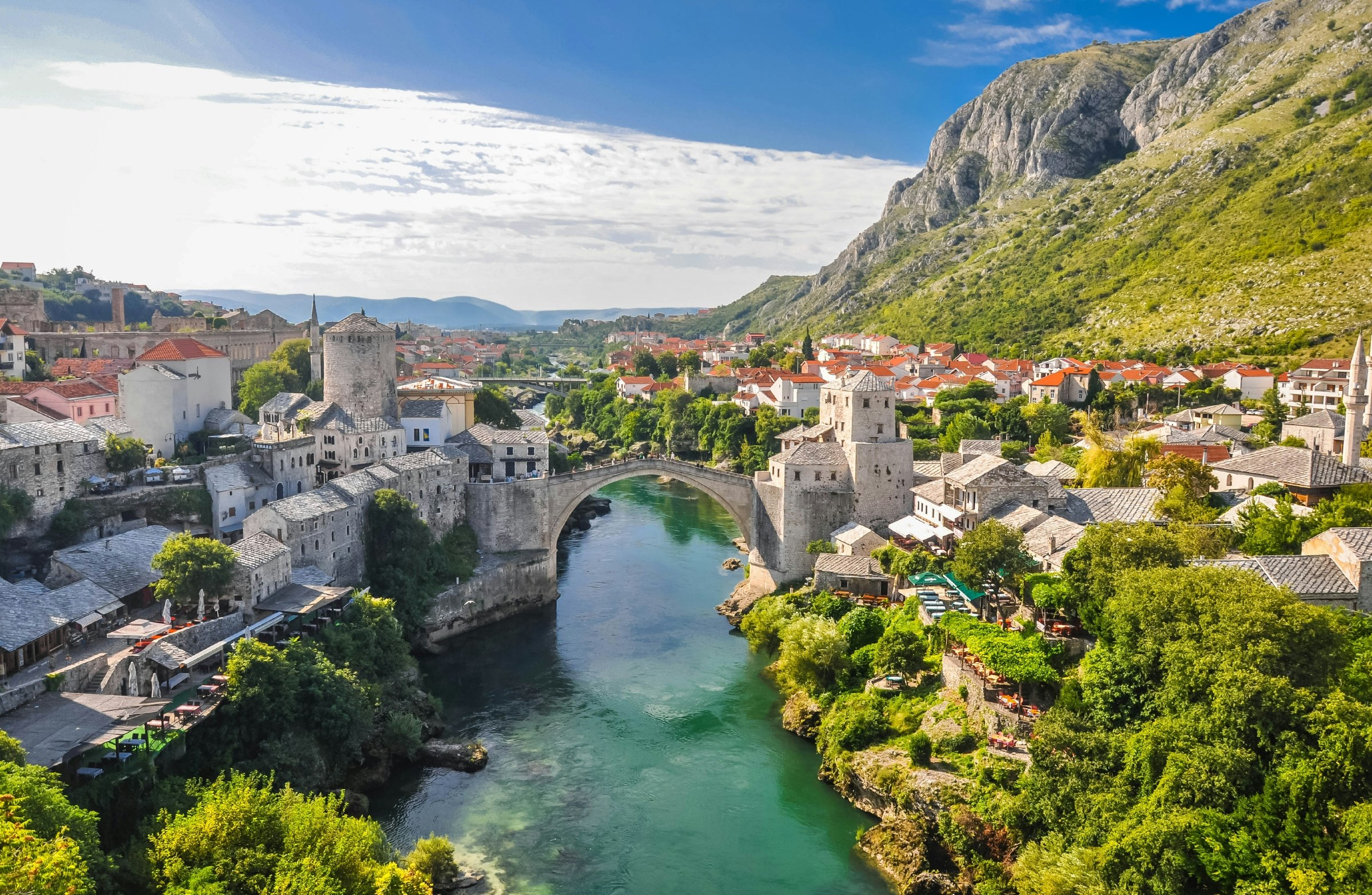 An aerial view of the city of Mostar in Bosnia and Hercegovina. The view shows the old bridge, which spans the river that runs through the city, and numerous white buildings surrounding it. In the background, green mountains are visible.