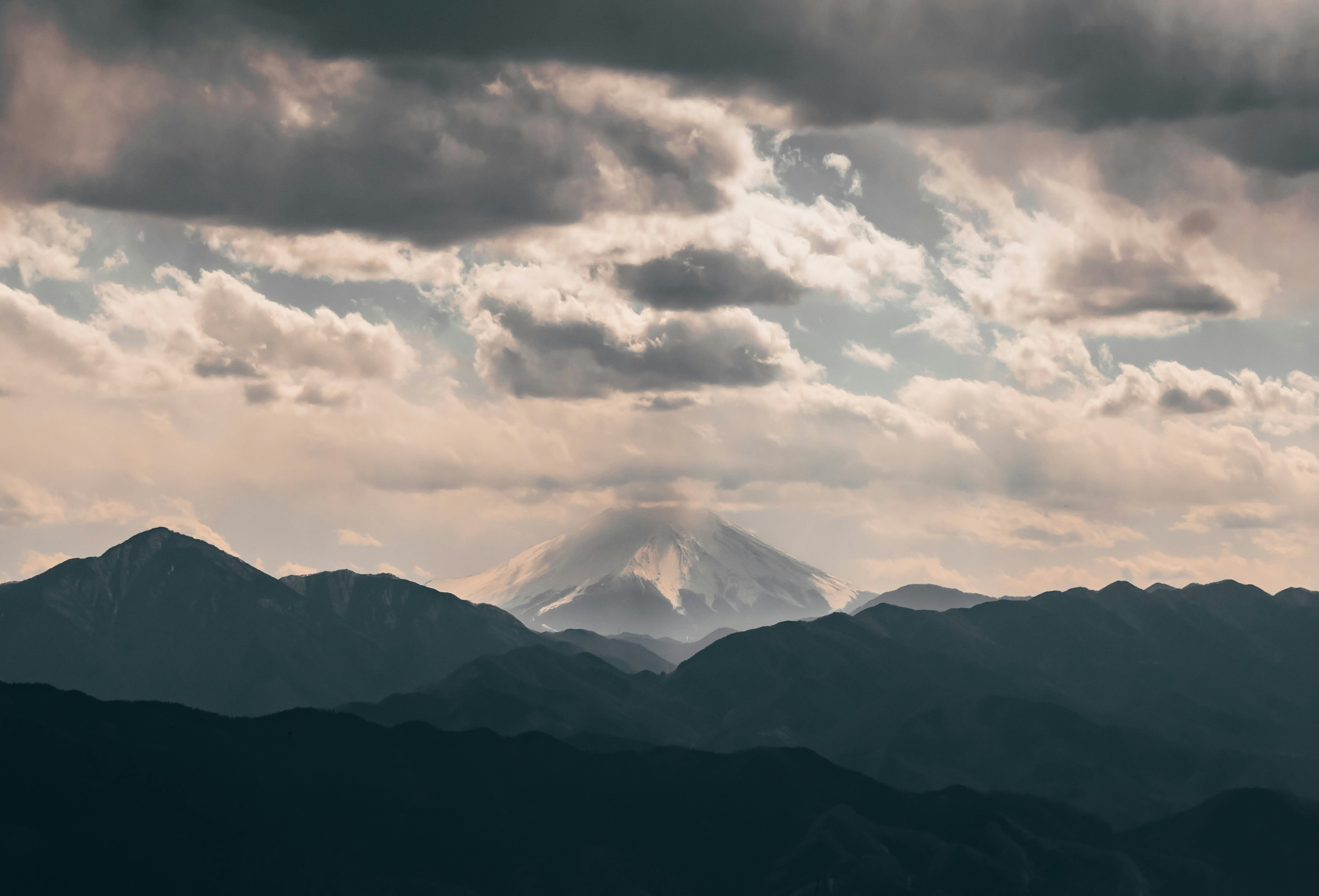 A distant view of Mt Fuji from Mt Takao. The iconic mountain rises above the surrounding hills and its snow-capped peak is enveloped by wispy white cloud.