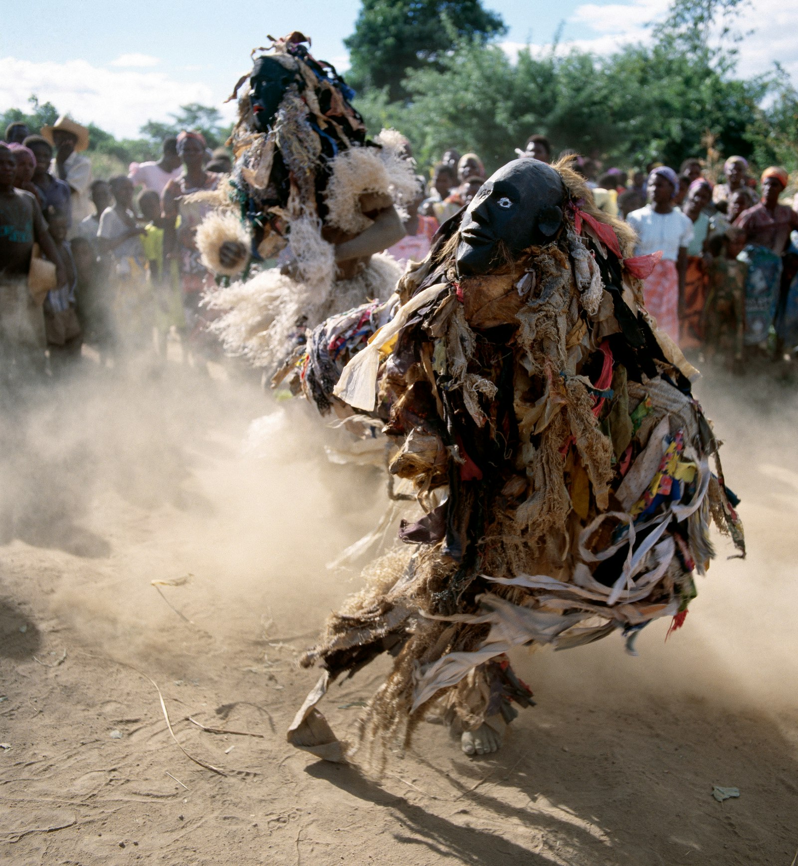 Two masked dancers, both clothed in countless shreds of hanging fabric, kick up dust as they perform in front of a crowd of Malawians in Mua