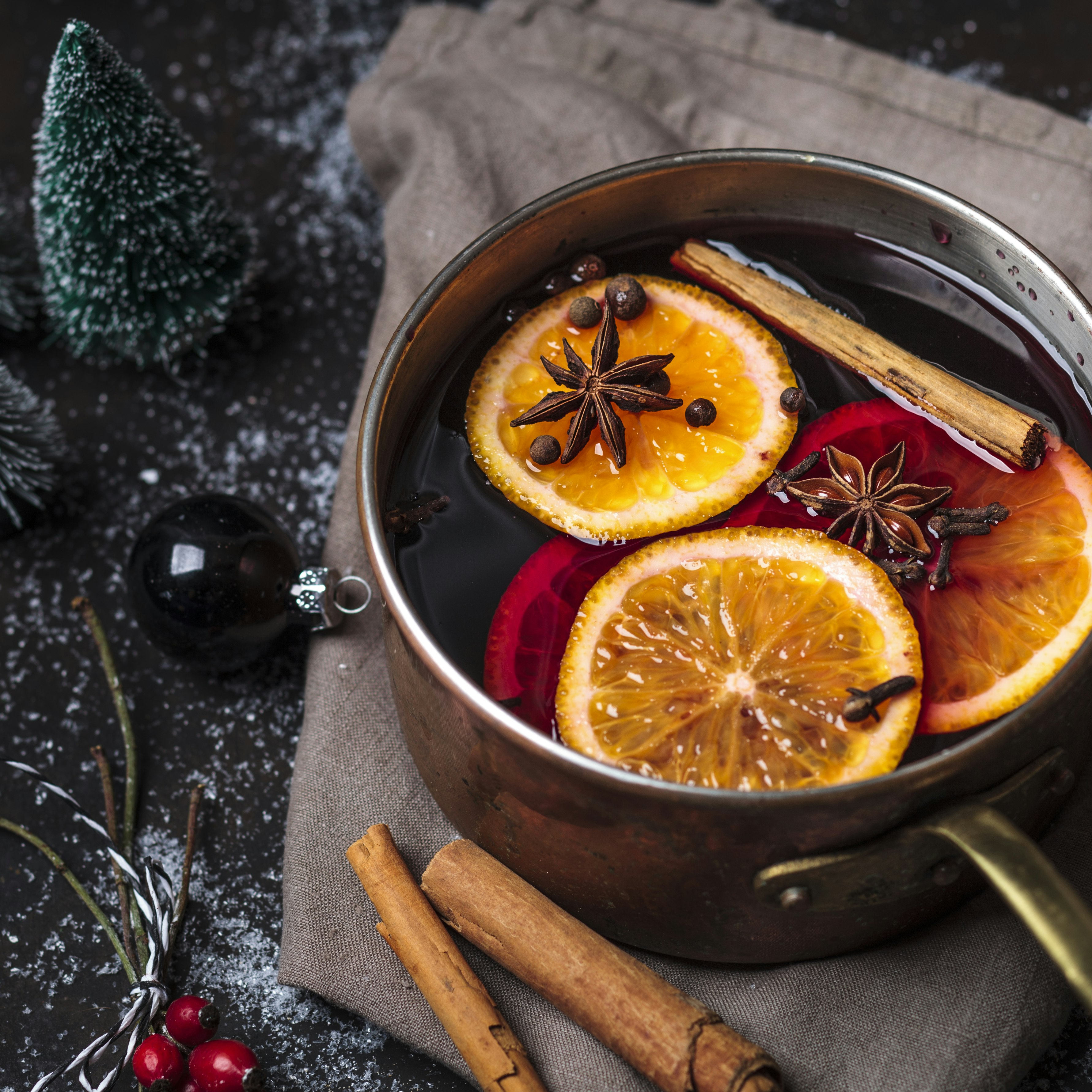 A silver pan filled with mulled wine, cinnamon sticks, orange slices and star anise is surrounded by miniature frosted Christmas trees, cranberries and an ornament