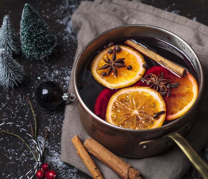 A silver pan filled with mulled wine, cinnamon sticks, orange slices and star anise is surrounded by miniature frosted Christmas trees, cranberries and an ornament