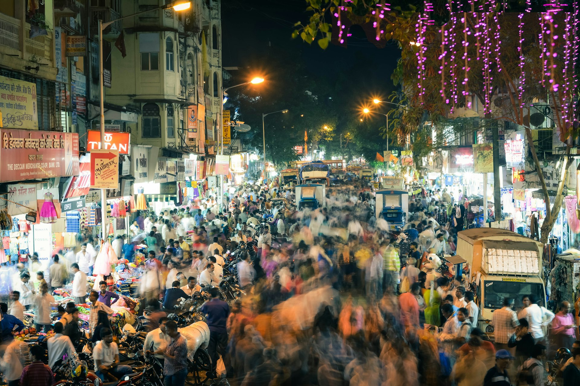 A view of a very busy road in Mumbai, flanked on either side by makeshift stalls and shops. The crowds walking through the street are blurred slightly, giving the appearance that they are in motion.